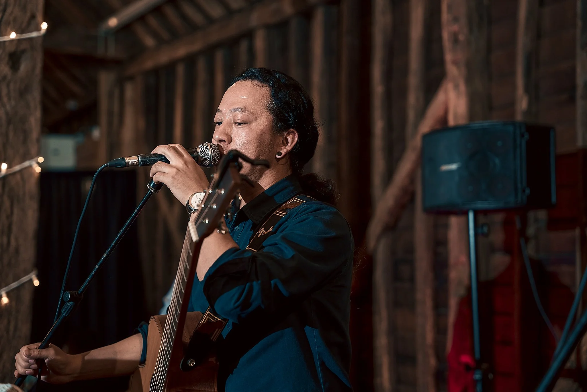 A man with black hair, wearing a black shirt, is singing into a microphone and playing an acoustic guitar in a rustic wooden setting.