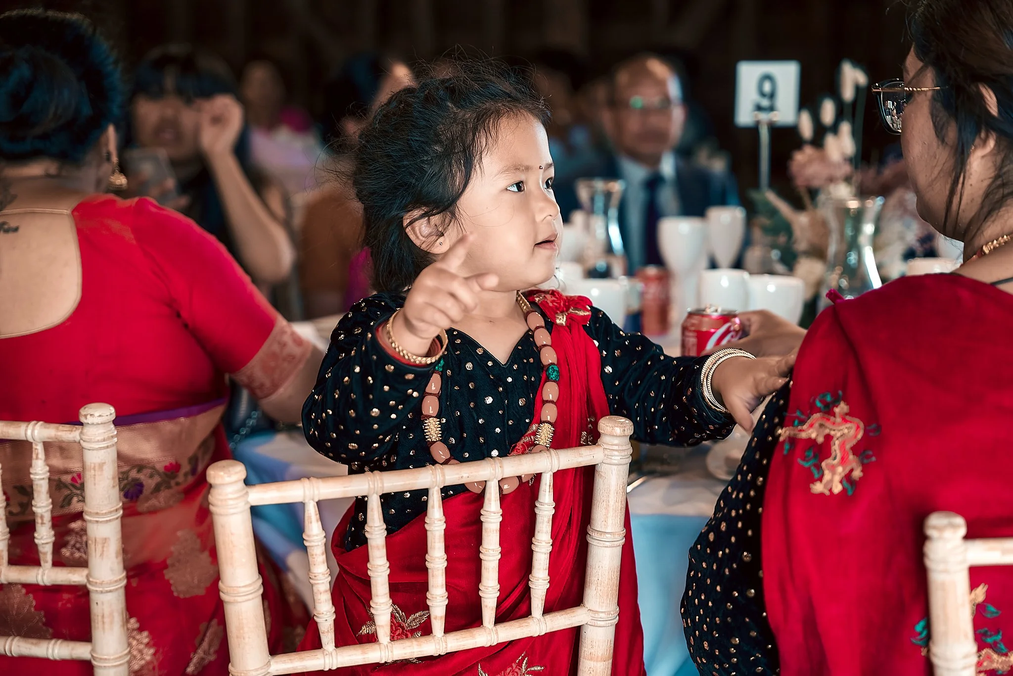 A young girl wearing traditional Indian attire and jewelry is sitting at a banquet table with other adults, pointing and engaging in conversation during a celebration or wedding reception.