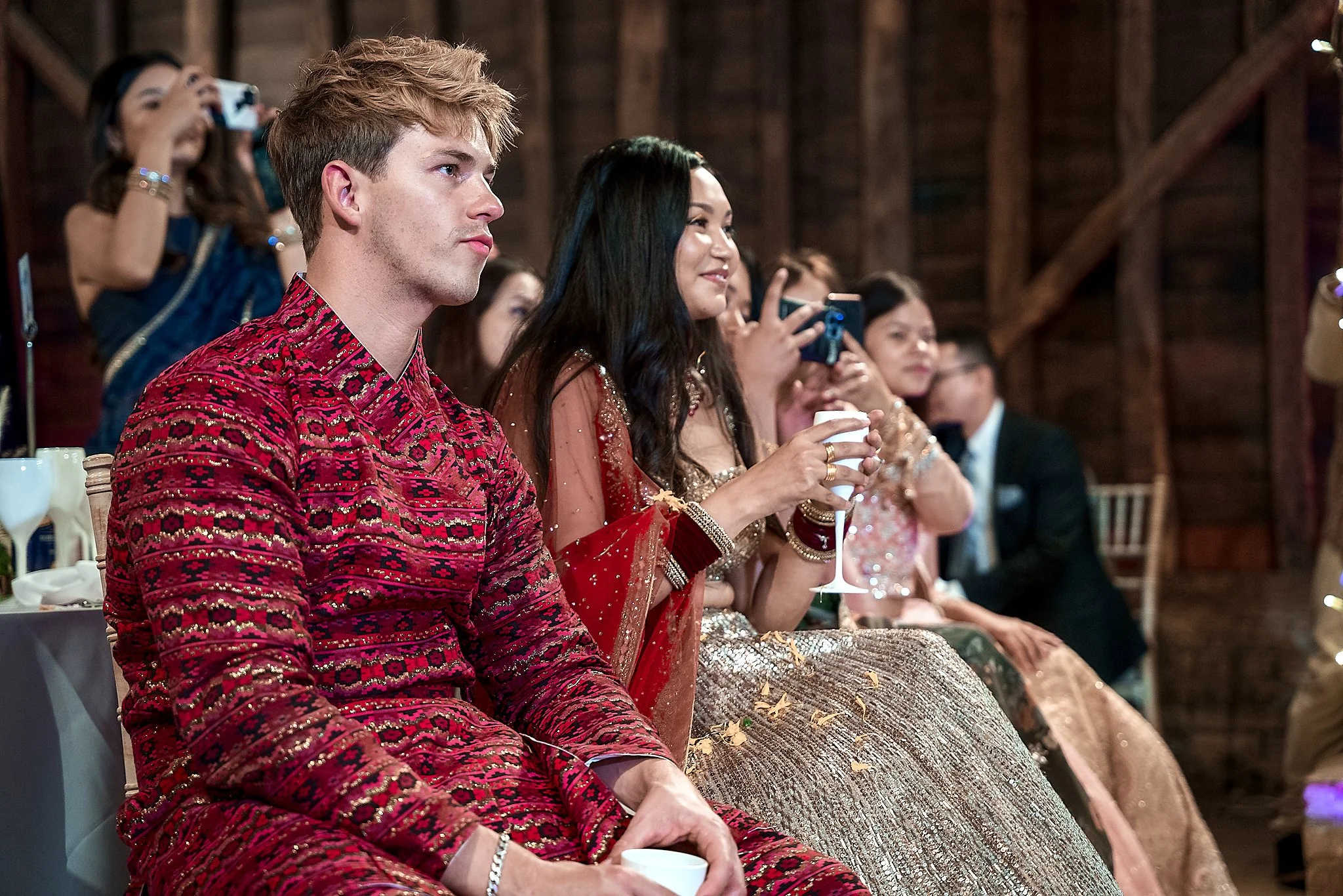 Group of people sitting at a wedding reception, dressed in traditional Indian attire, with some watching and others taking photos.