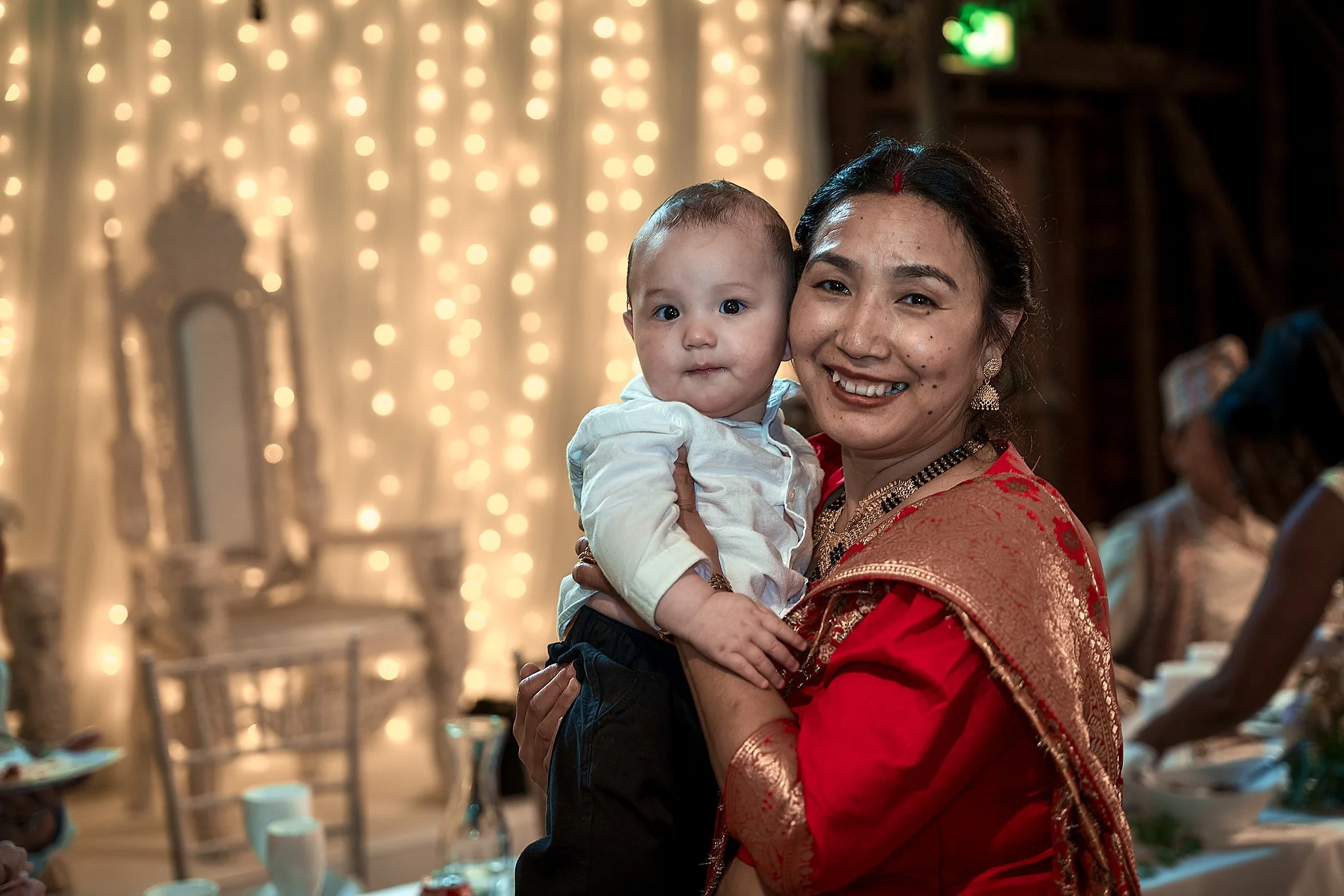 A smiling woman holding a baby in a festive indoor setting with warm lights and elegant decor in the background.