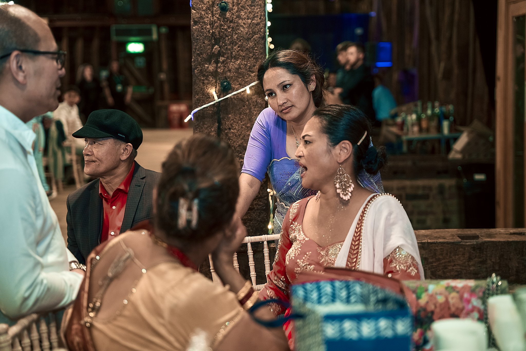 A group of people gathered at a social event, with women dressed in traditional Indian attire engaged in conversation, and men in the background.