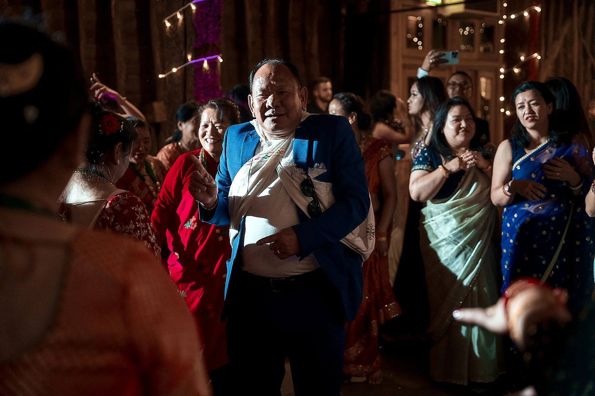 A man in a blue blazer dancing at a crowded party with people in colorful traditional Indian attire around him.