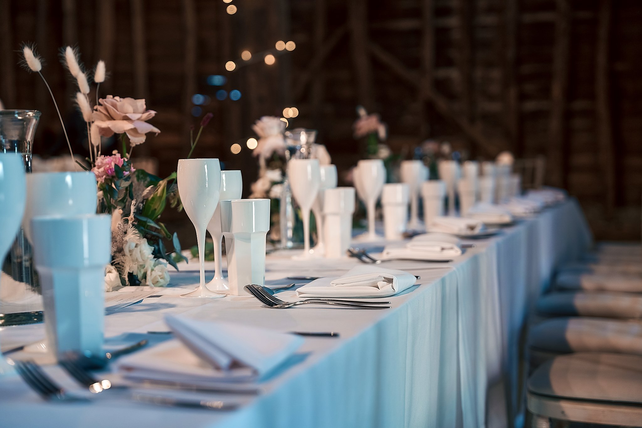 Elegant banquet table setup with white tablecloth, floral arrangements, white plates, silverware, and glasses in a rustic wooden venue.