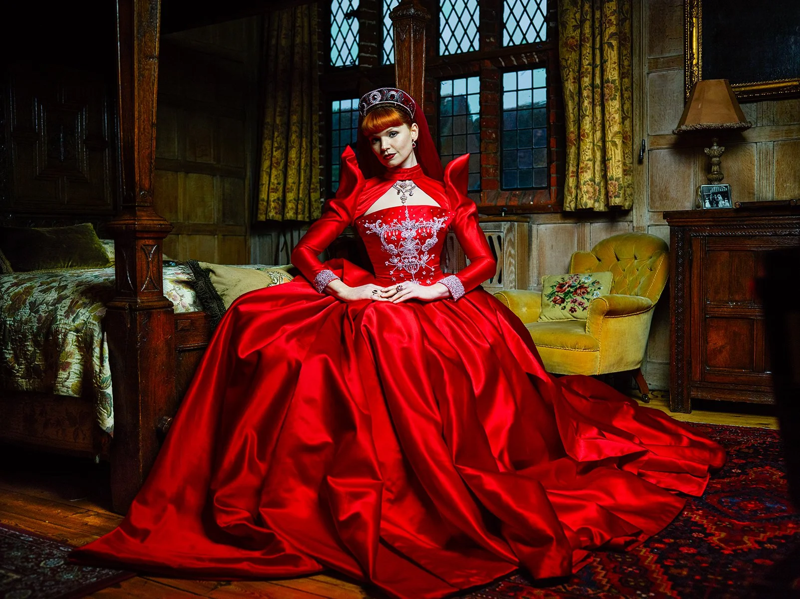 A woman dressed in an elaborate red gown with intricate white embroidery, sitting on a bed in a vintage-style room with dark wood paneling, large windows, and yellow upholstered chairs.