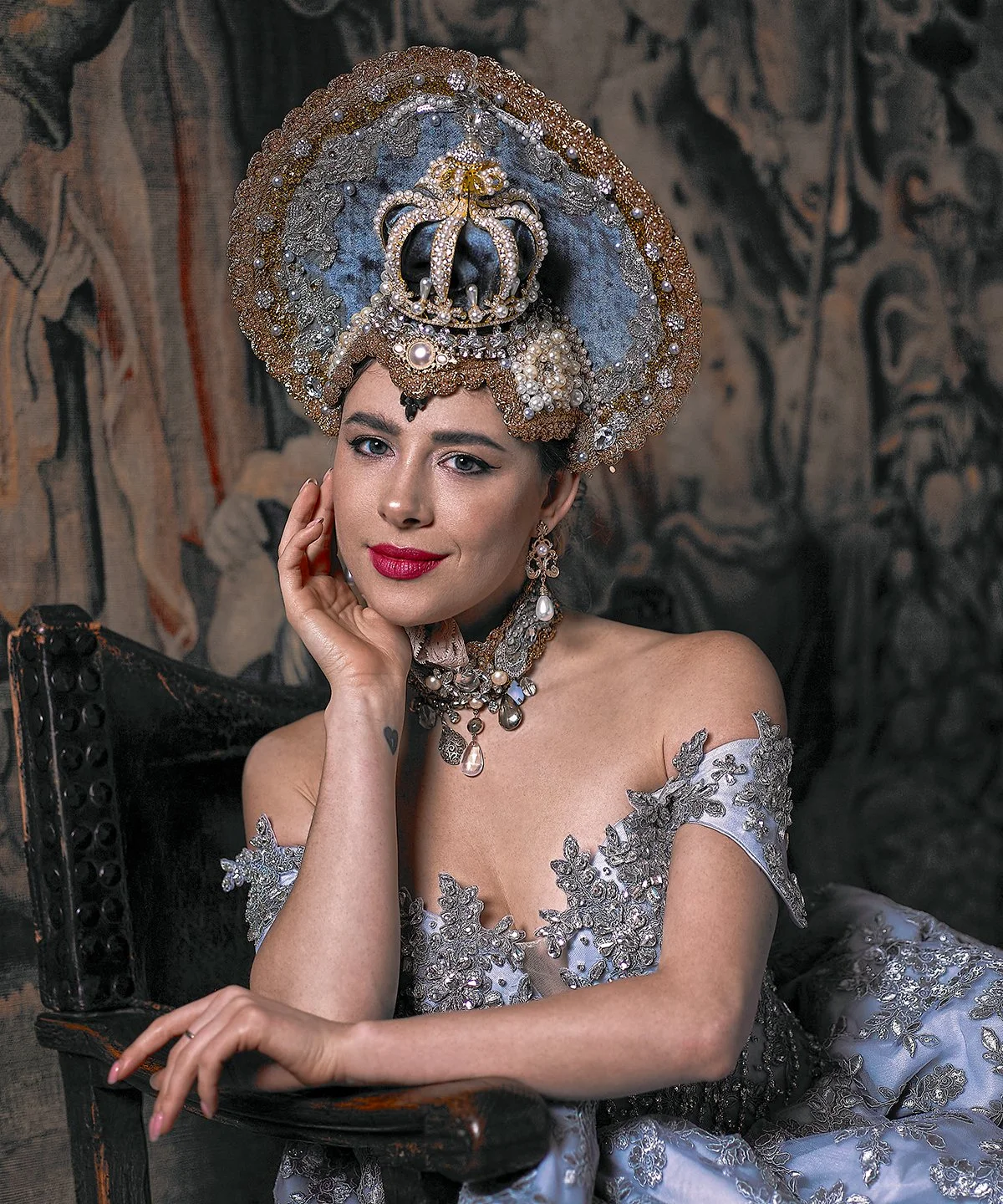 A woman wearing an elaborate crown and ornate jewelry, dressed in a silver embroidered gown, sitting on a dark wooden chair against a tapestry background.