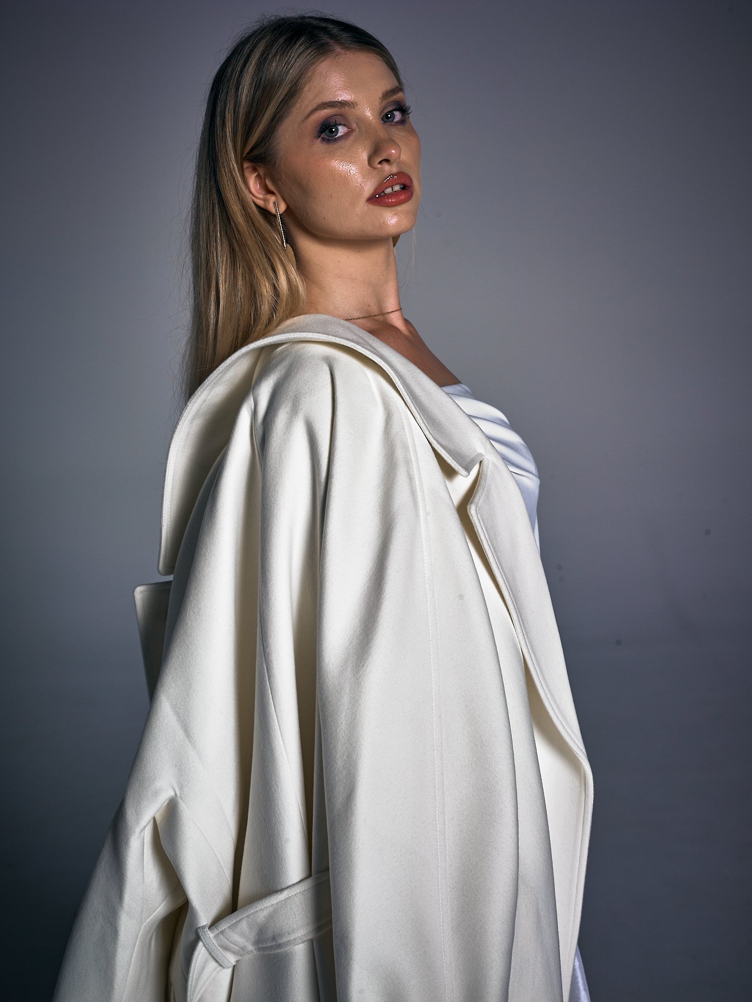 Portrait of a young woman with long blonde hair, wearing a cream-colored coat draped over her shoulders, and looking over her shoulder.