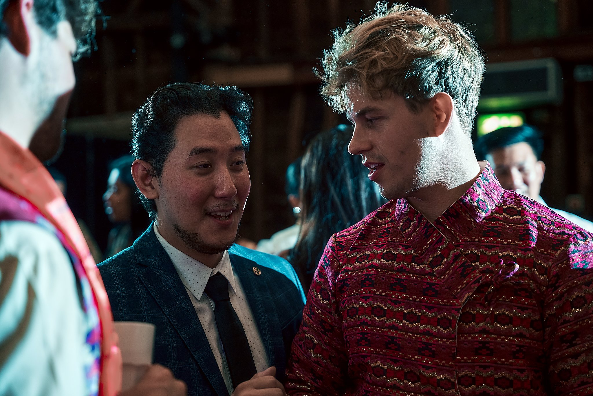 Two men having a conversation at a social event, one in a checked suit and the other in a patterned shirt, with blurred people in the background.