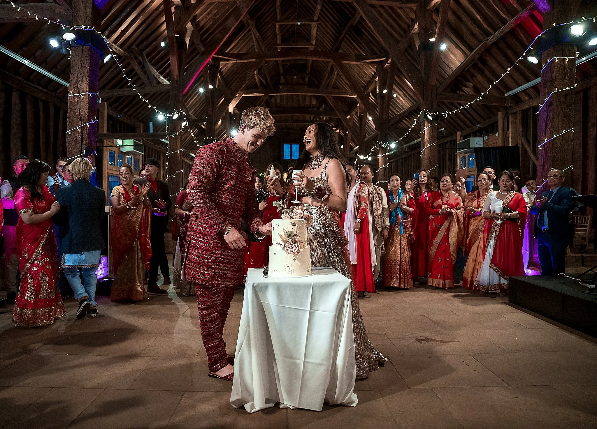 A celebration scene of a wedding reception with the bride and groom cutting the cake, surrounded by many guests dressed in colorful traditional attire inside a rustic wooden hall decorated with string lights.