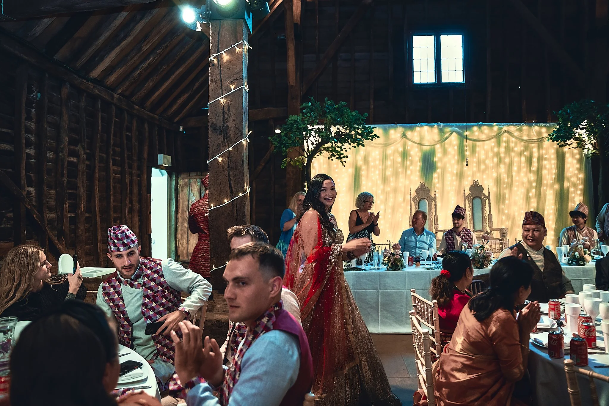 People gathered at a celebration in a rustic barn with string lights and a backdrop of curtains with fairy lights. Some guests are dressed in traditional South Asian attire, and a woman in a red and gold lehenga is smiling in the center.
