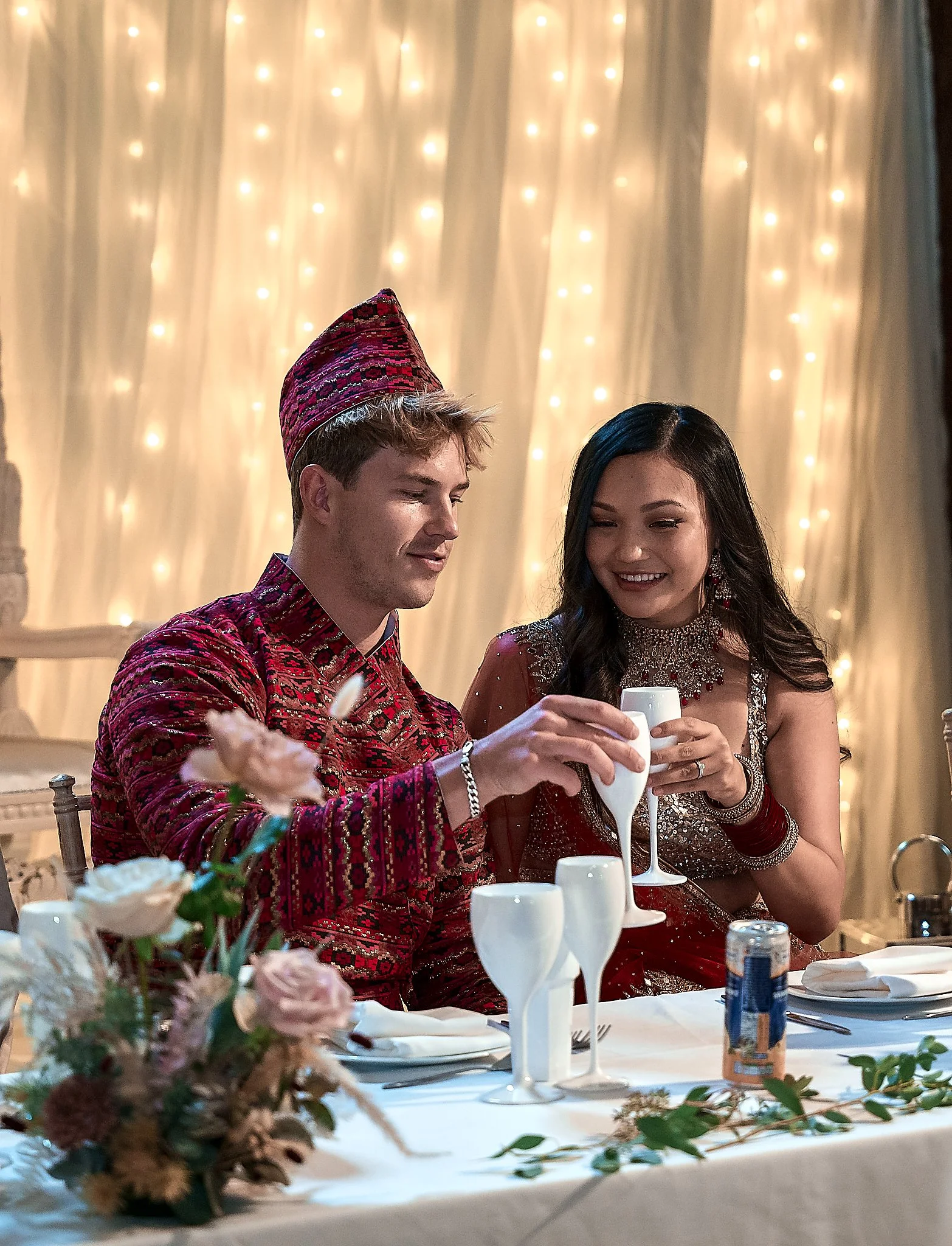 A man and woman at a decorated dinner table raising glasses in a toast, with fairy lights in the background.