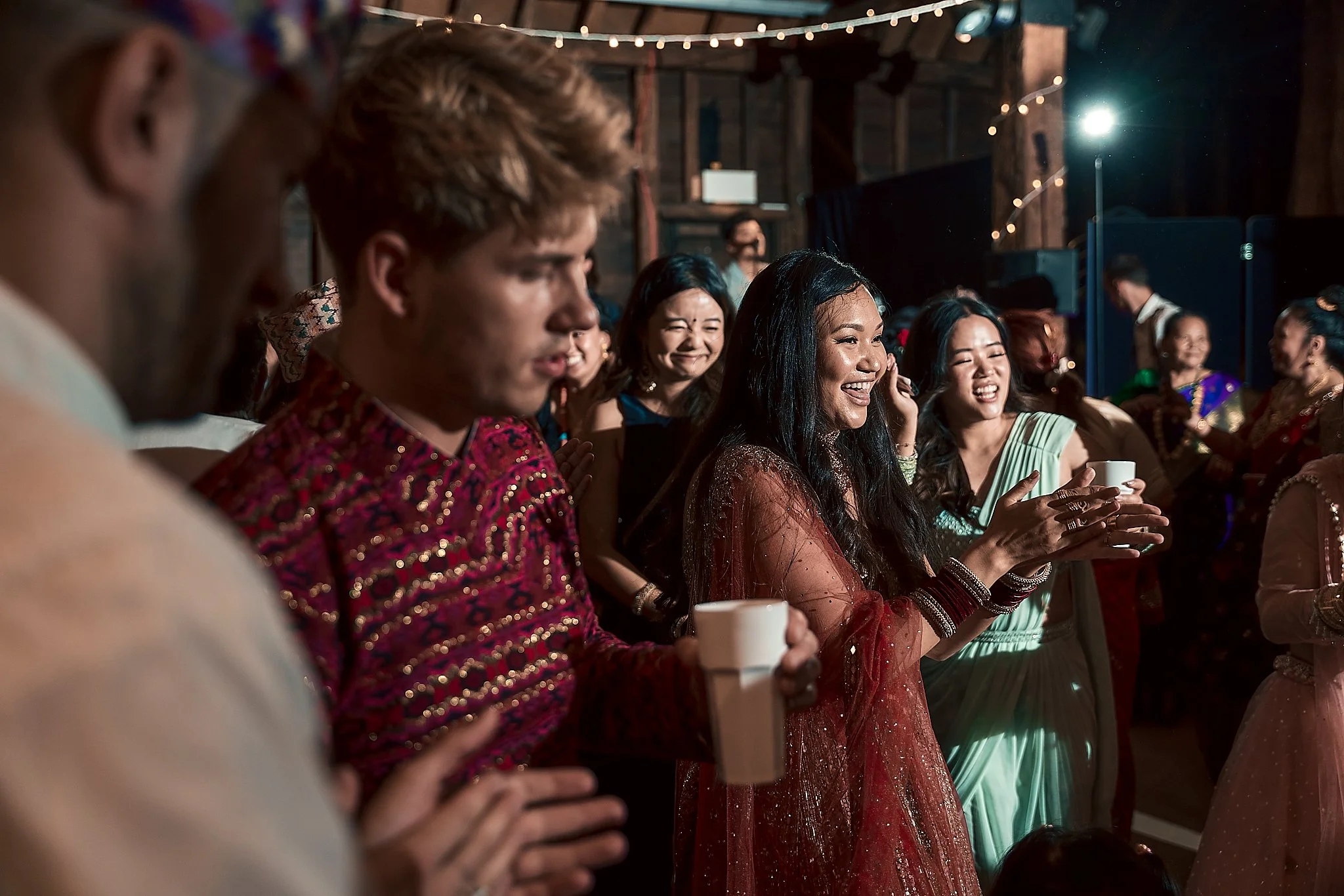Group of people dressed in colorful traditional and modern attire, enjoying a celebration or event in a decorated indoor setting with string lights.