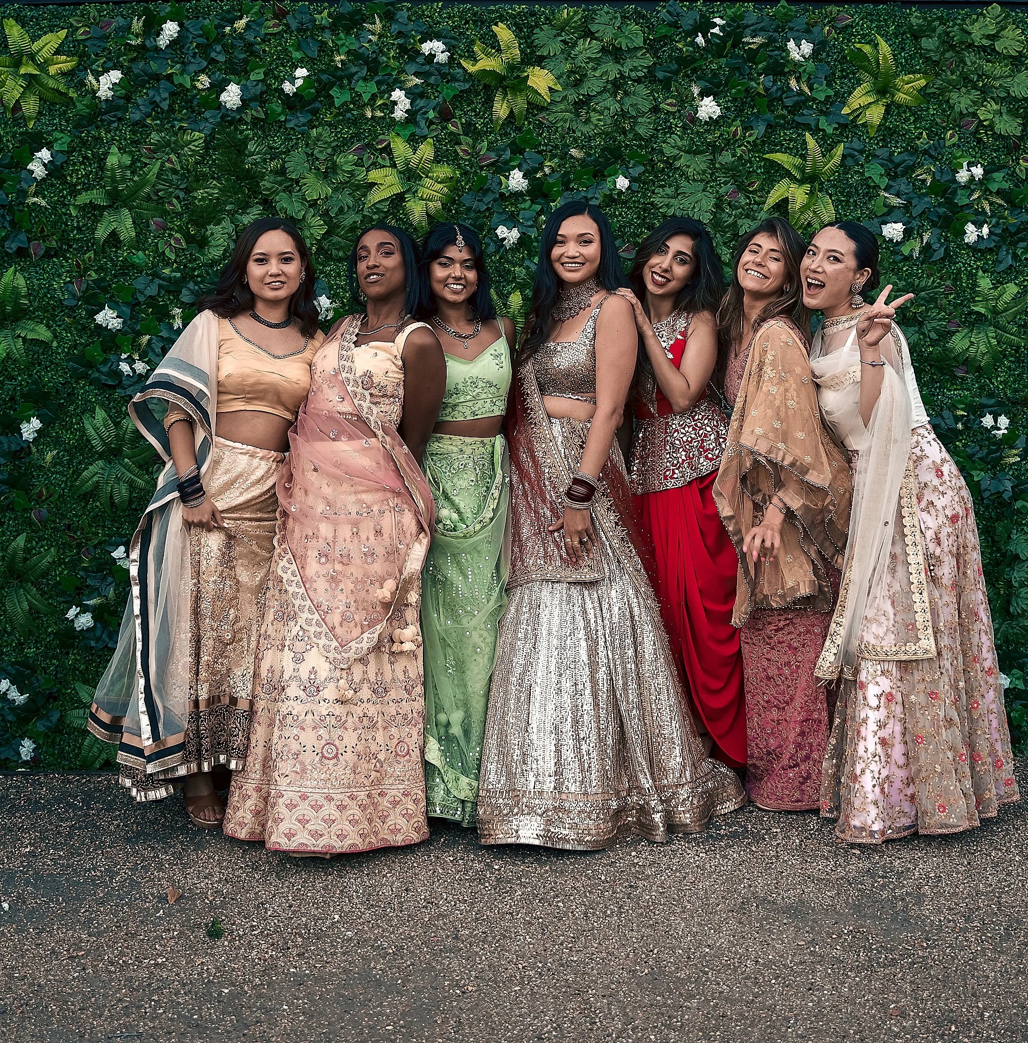 Group of women dressed in traditional Indian clothing posing in front of a green leafy backdrop at a celebration.