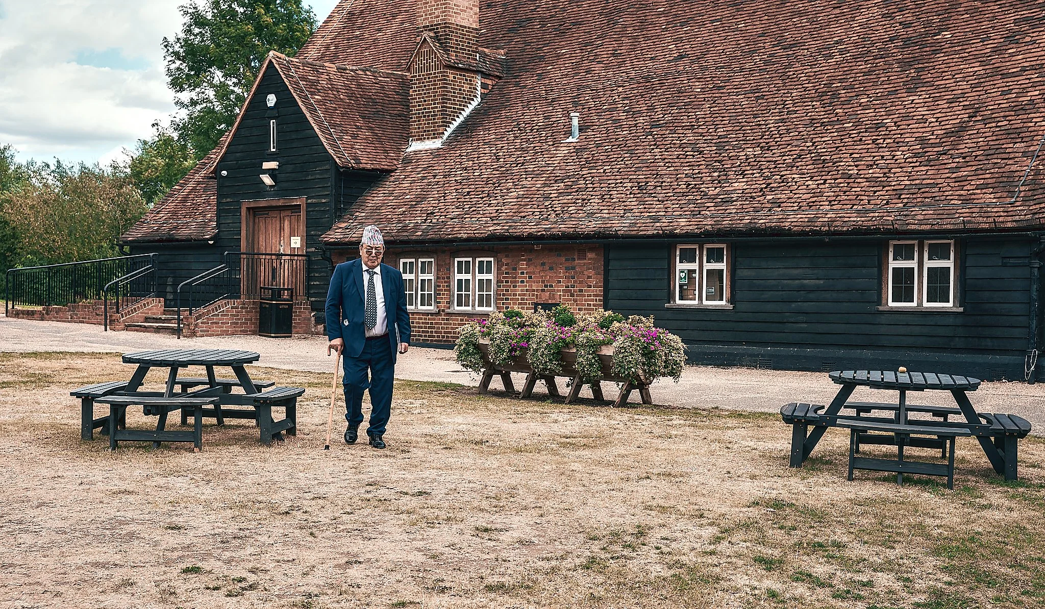 An elderly man in a suit and tie walks with a cane in front of a black wooden building with a red-tiled roof. There are pink and white flowers in large planters and several black picnic tables on the lawn.