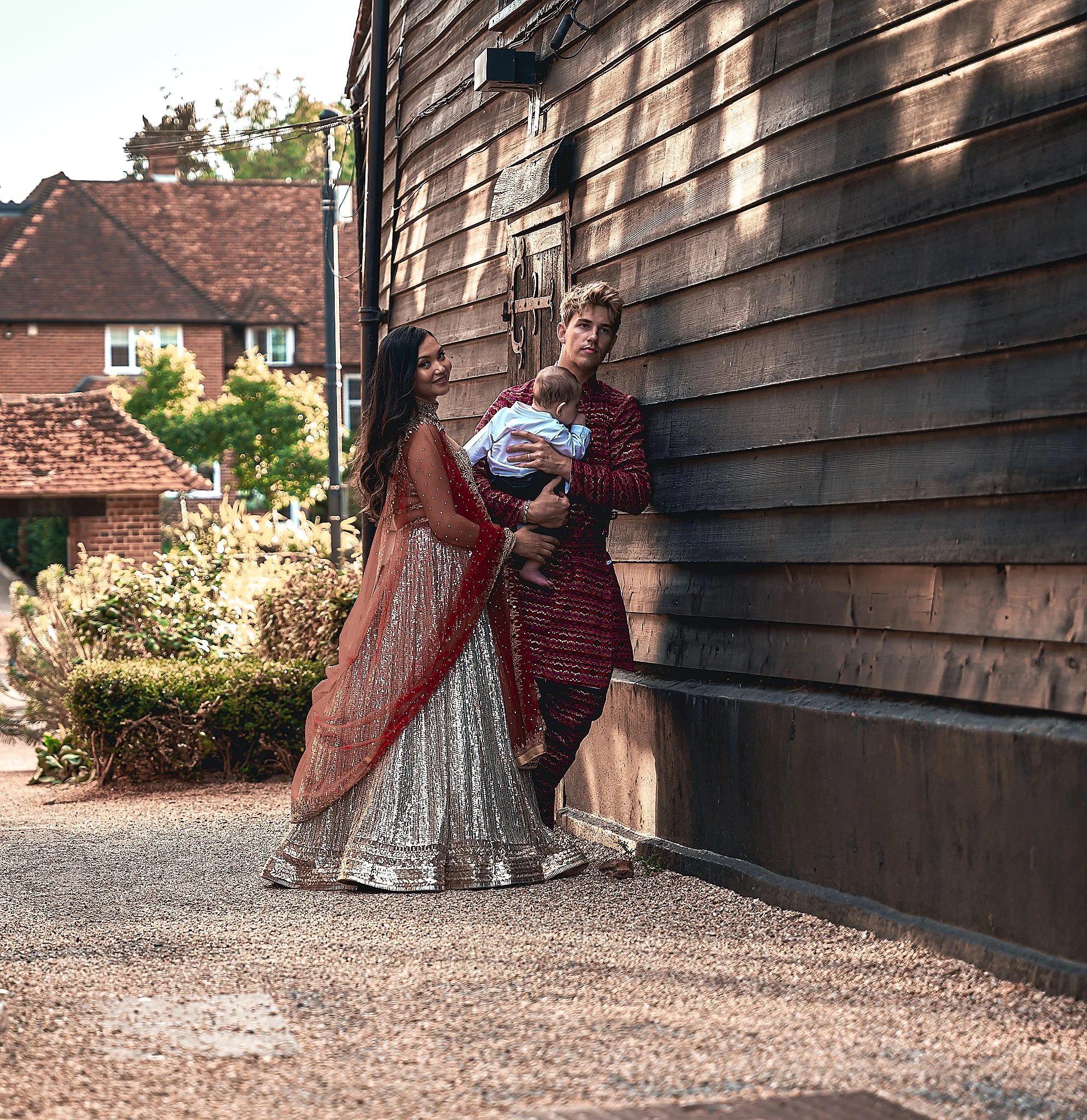 A couple dressed in traditional South Asian attire standing outside a wooden building with a baby, man holding the baby and woman smiling.