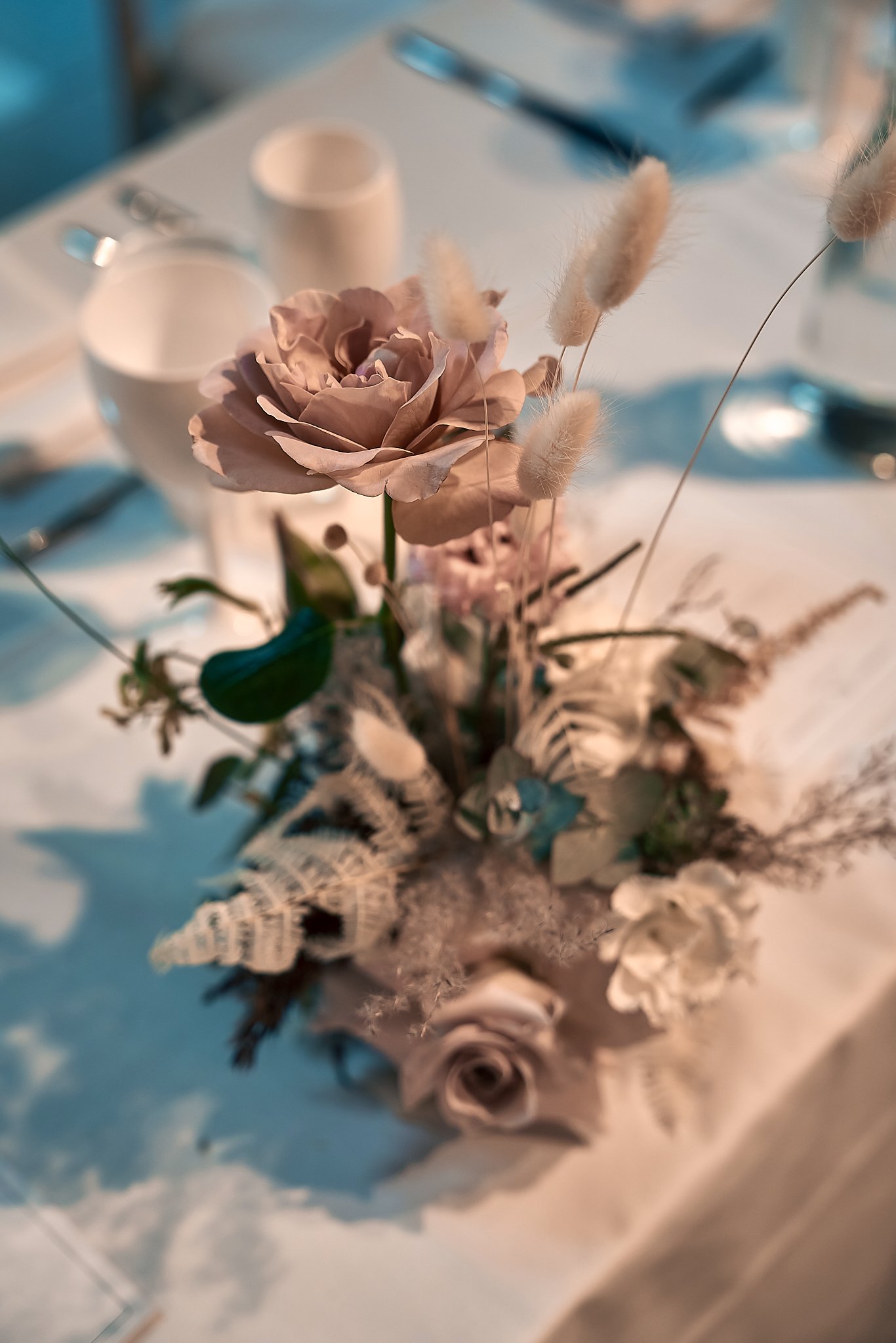 A floral centerpiece with pink roses, beige dried flowers, and greenery on a table at a formal event.