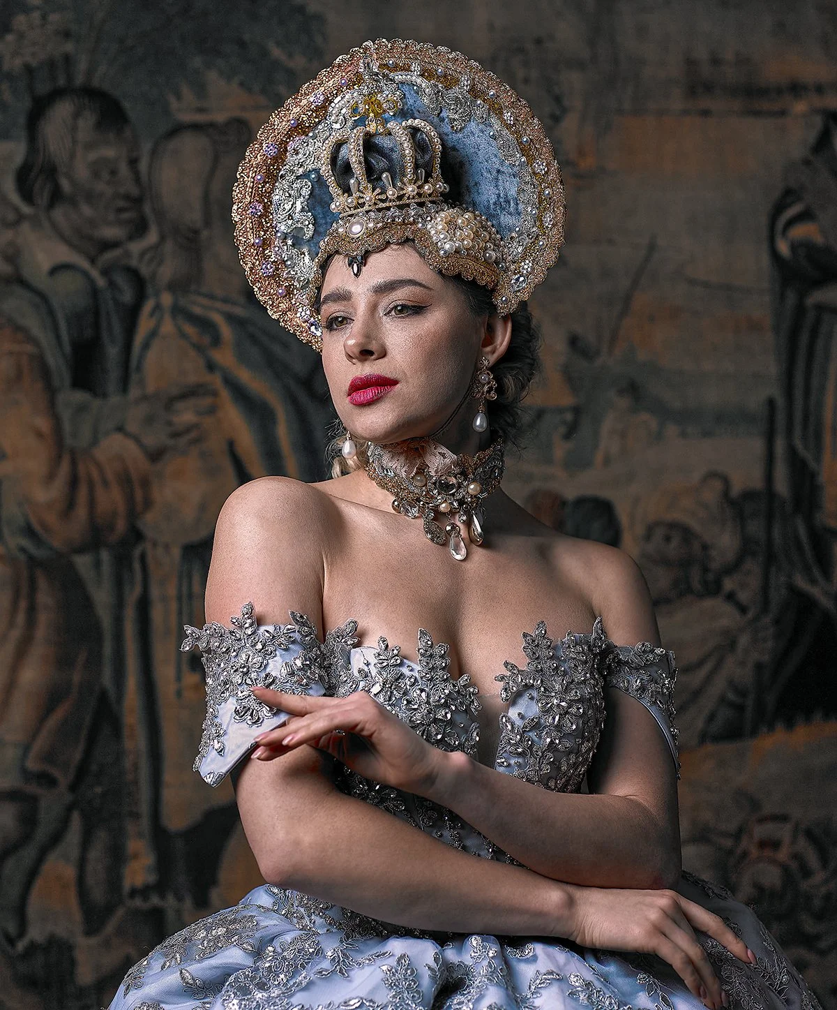 A woman in an ornate silver and pearl costume with a large jeweled crown, earrings, and necklace, standing in front of an art mural background.