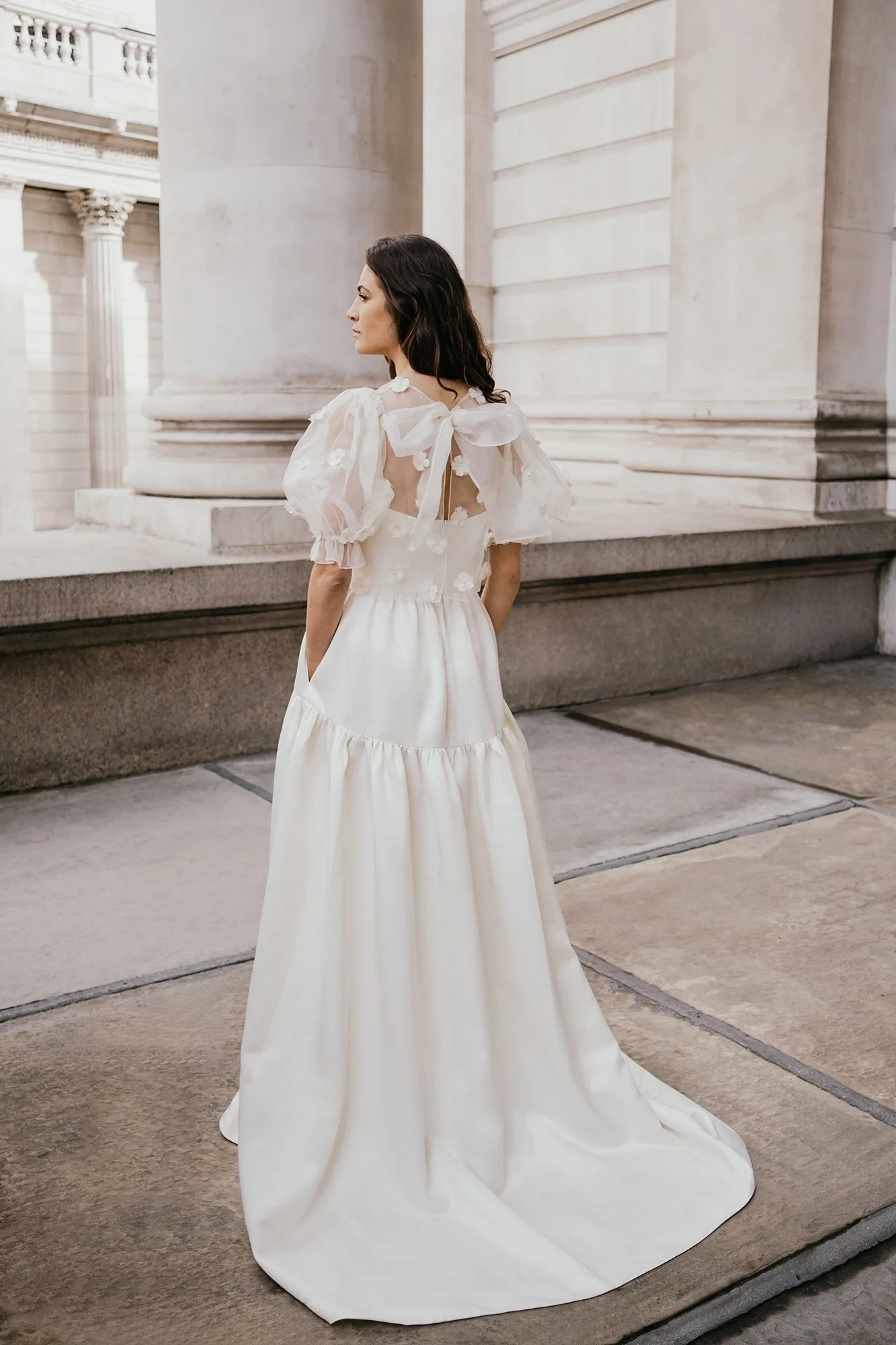 A woman in a white wedding dress with puffy sleeves and a large bow on the back, standing outside in front of a classical building with columns.