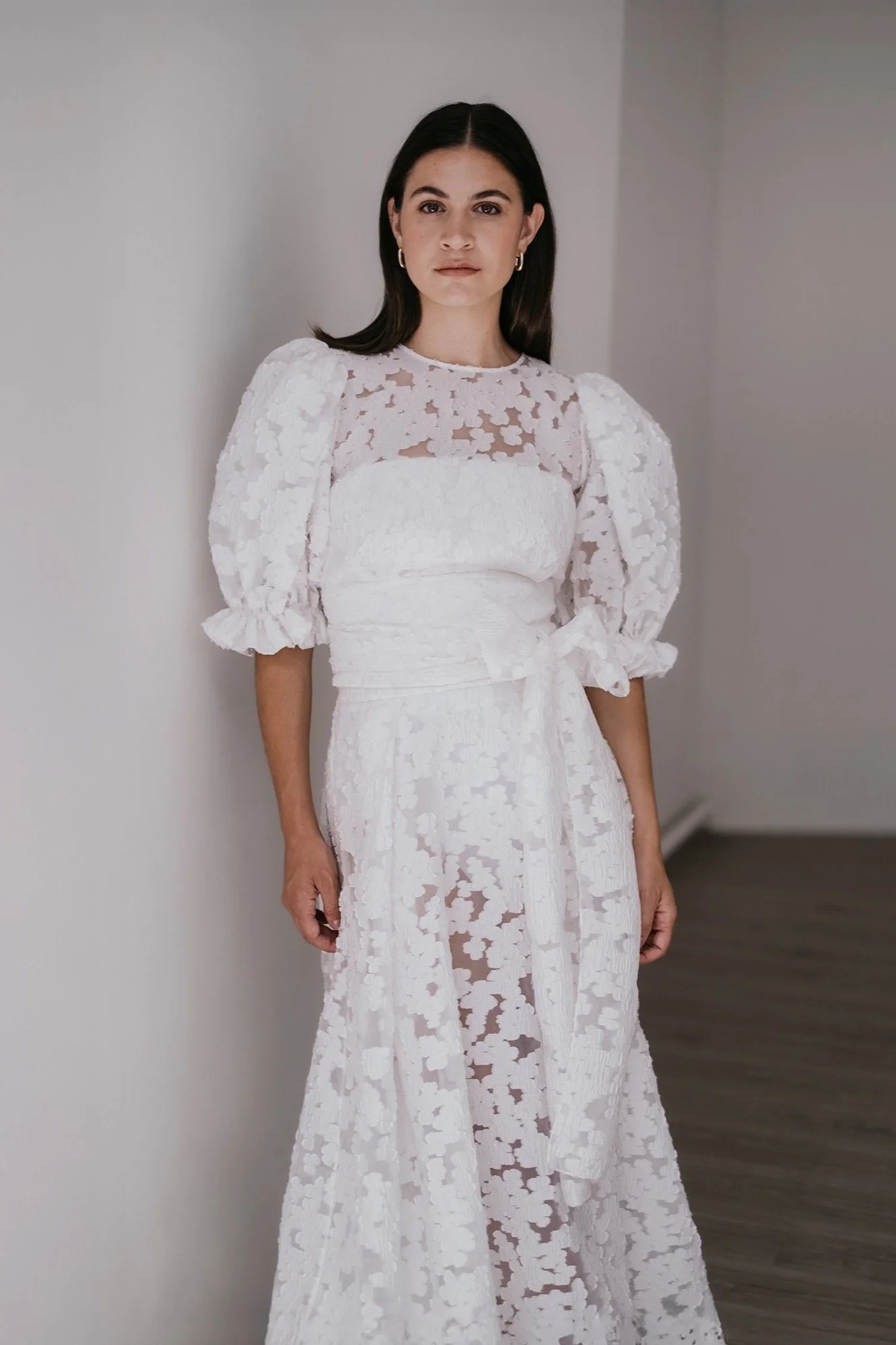 A woman standing indoors, wearing a white lace dress with puffed sleeves and a sheer lace top.