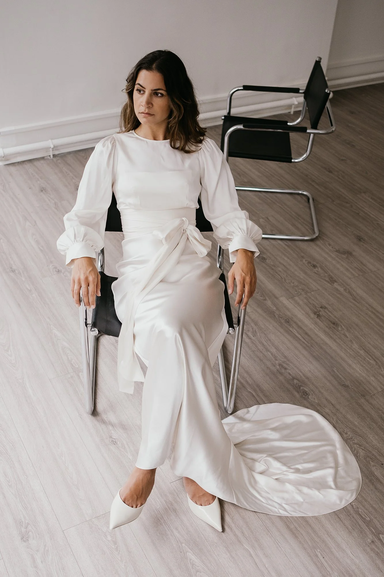 A woman in a white dress and pointed heels sitting in a black chair on a wooden floor, with an empty black chair behind her.