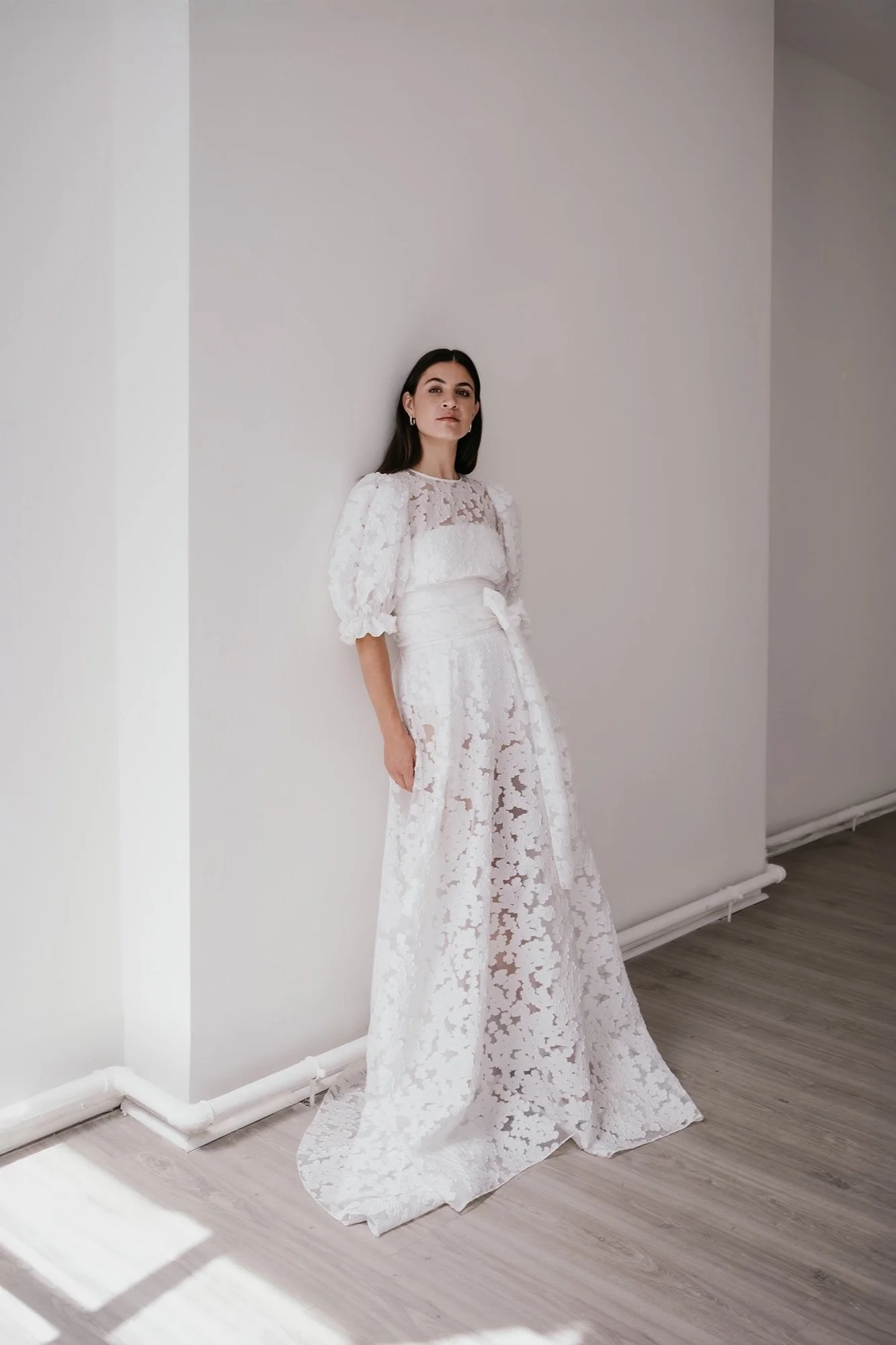A woman with dark hair wearing a white lace wedding dress with puffed sleeves, standing against a plain white wall in a minimalist room.