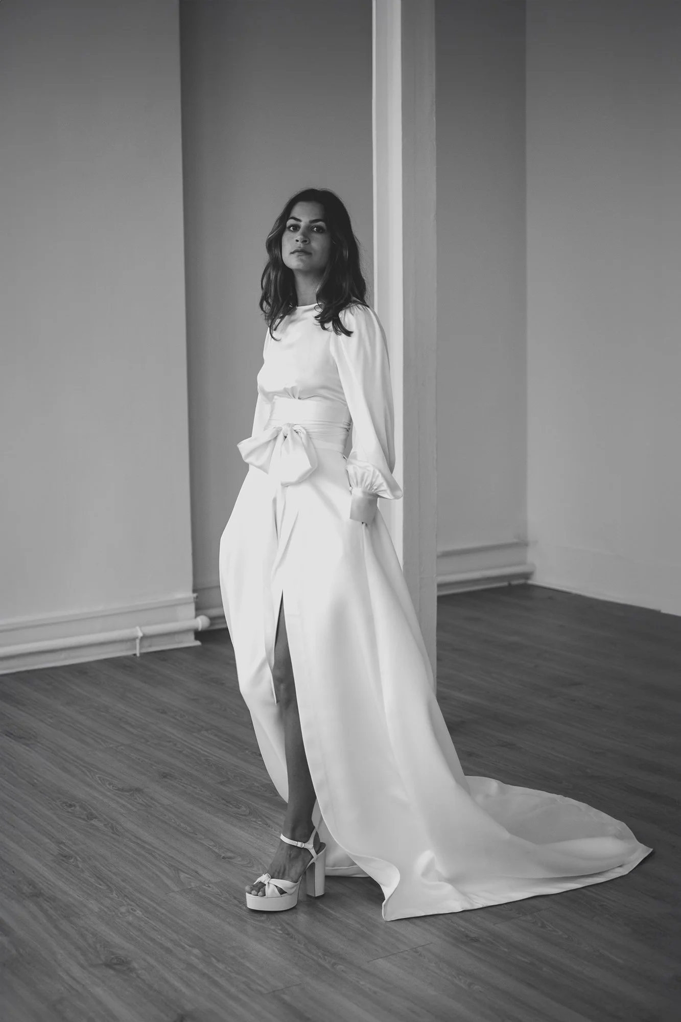 A woman in a long white dress with puffy sleeves and a large bow at the waist, standing on a wooden floor in a room with plain walls, wearing high platform shoes, looking at the camera.