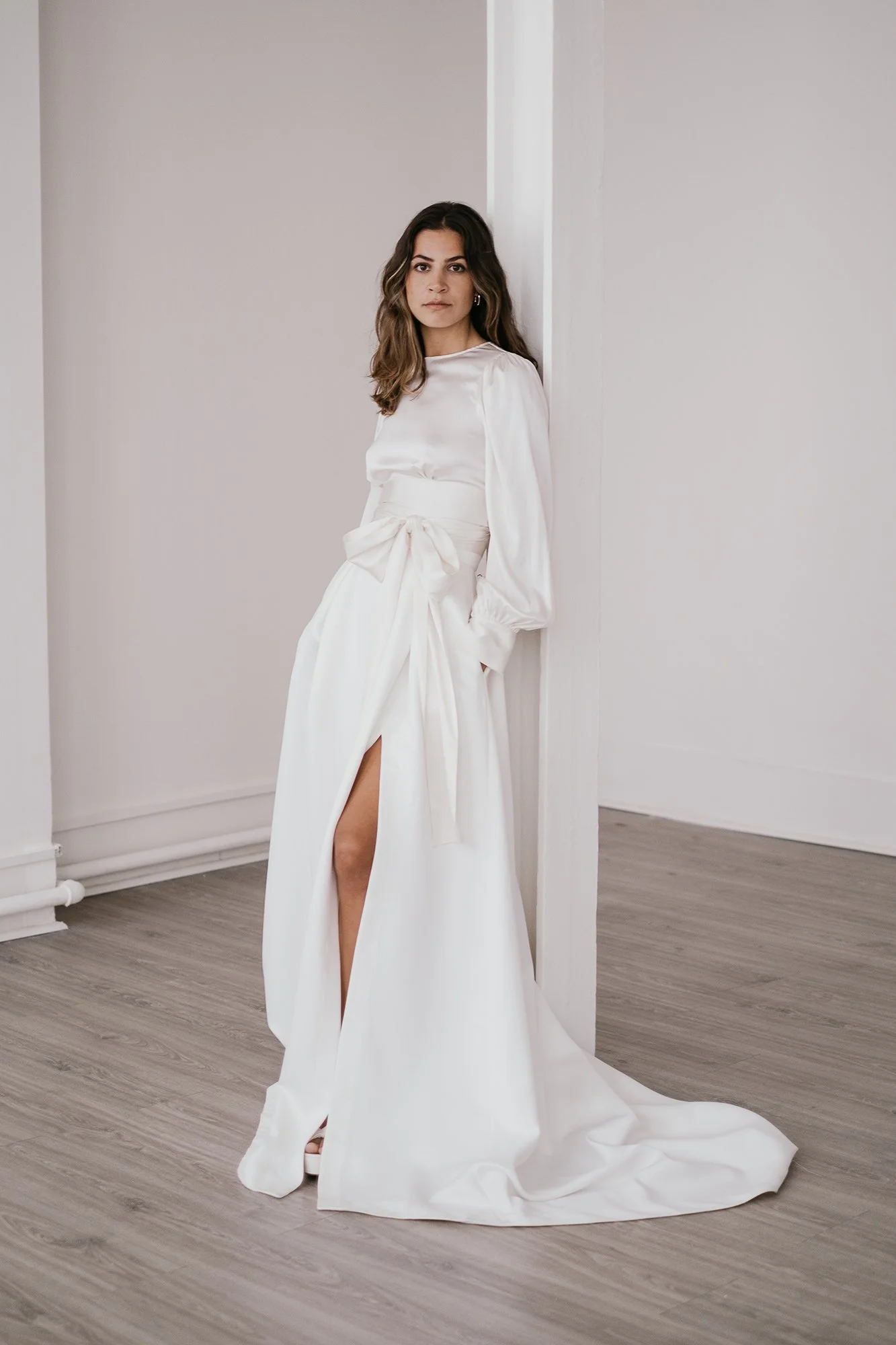 A woman in a white bridal gown with long sleeves, standing against a corner with a slit showing her leg, on a wooden floor in a minimalist room.
