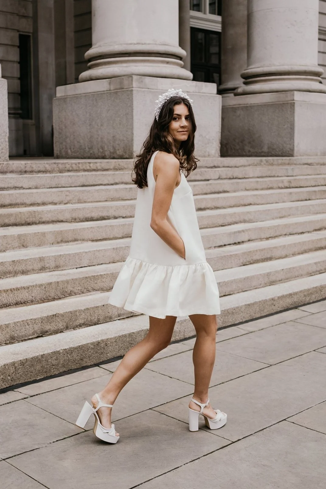 Young woman in white dress and heels standing on city steps with columns in the background.
