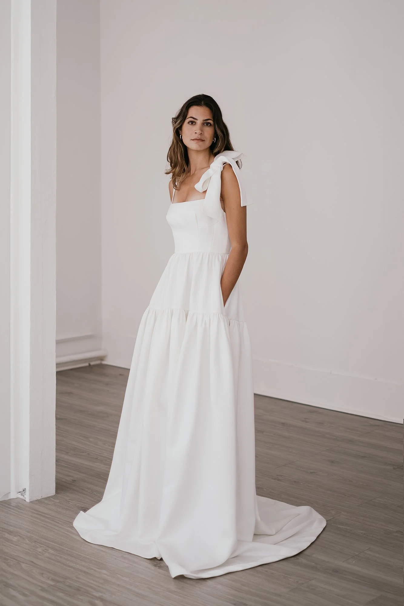 A woman in a white dress standing in a simple, minimalist room with wooden flooring and white walls.