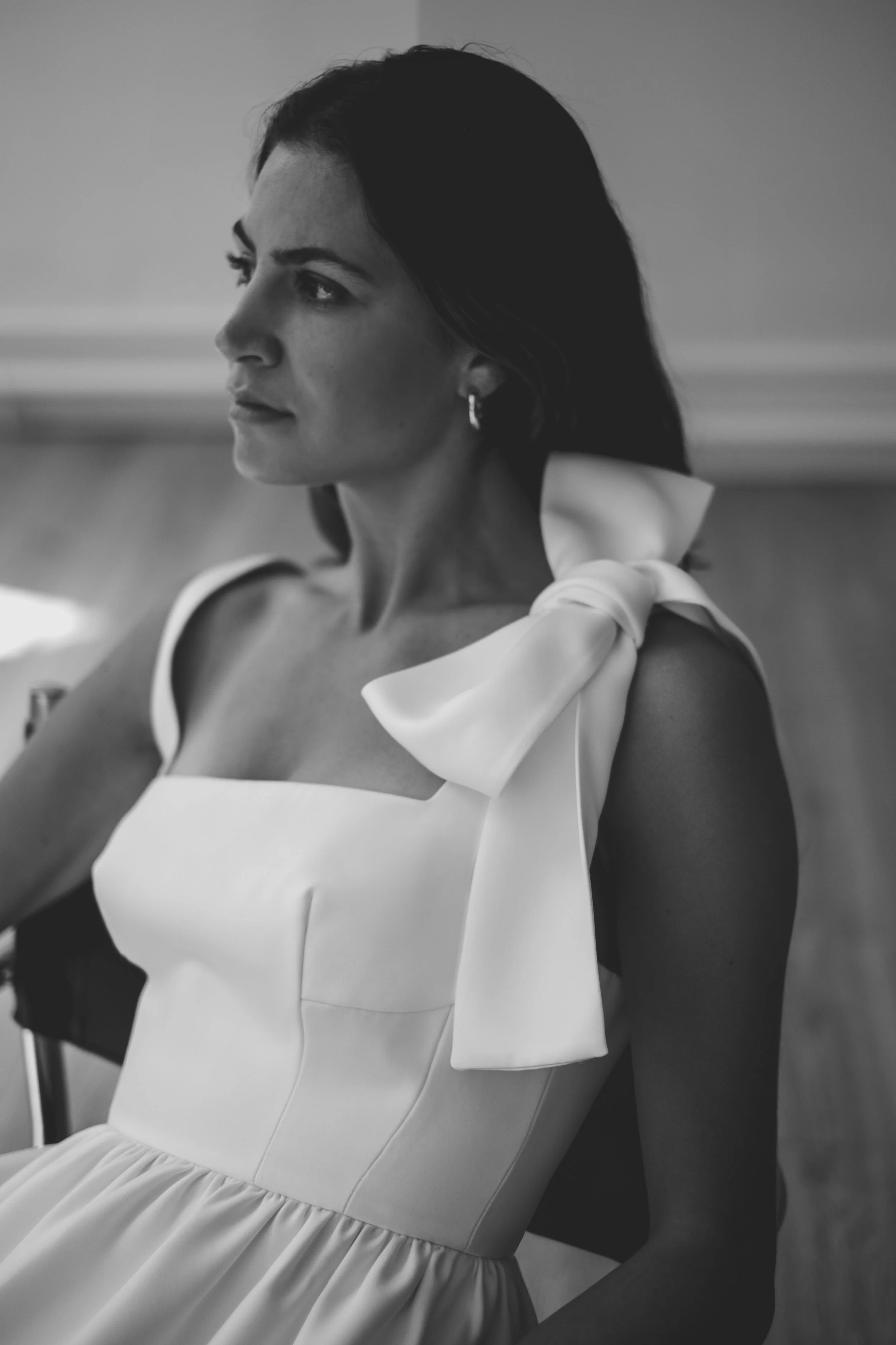 A black-and-white photograph of a woman with dark hair, dressed in a sleeveless gown with a large bow on her shoulder, sitting indoors with a thoughtful expression.