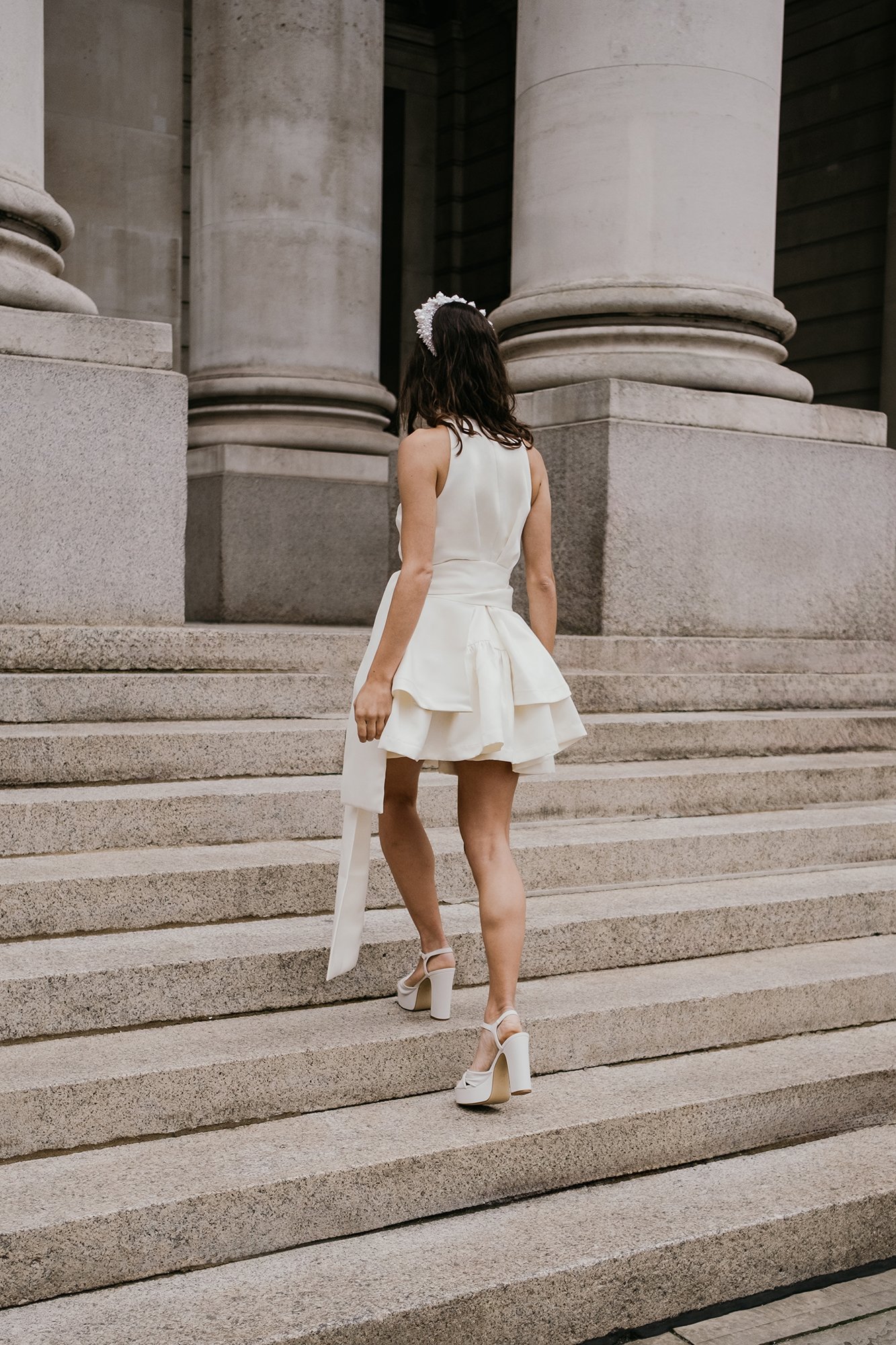 A woman wearing a white dress with a flared skirt and high-heeled shoes walking up stone steps outside a building with large columns.