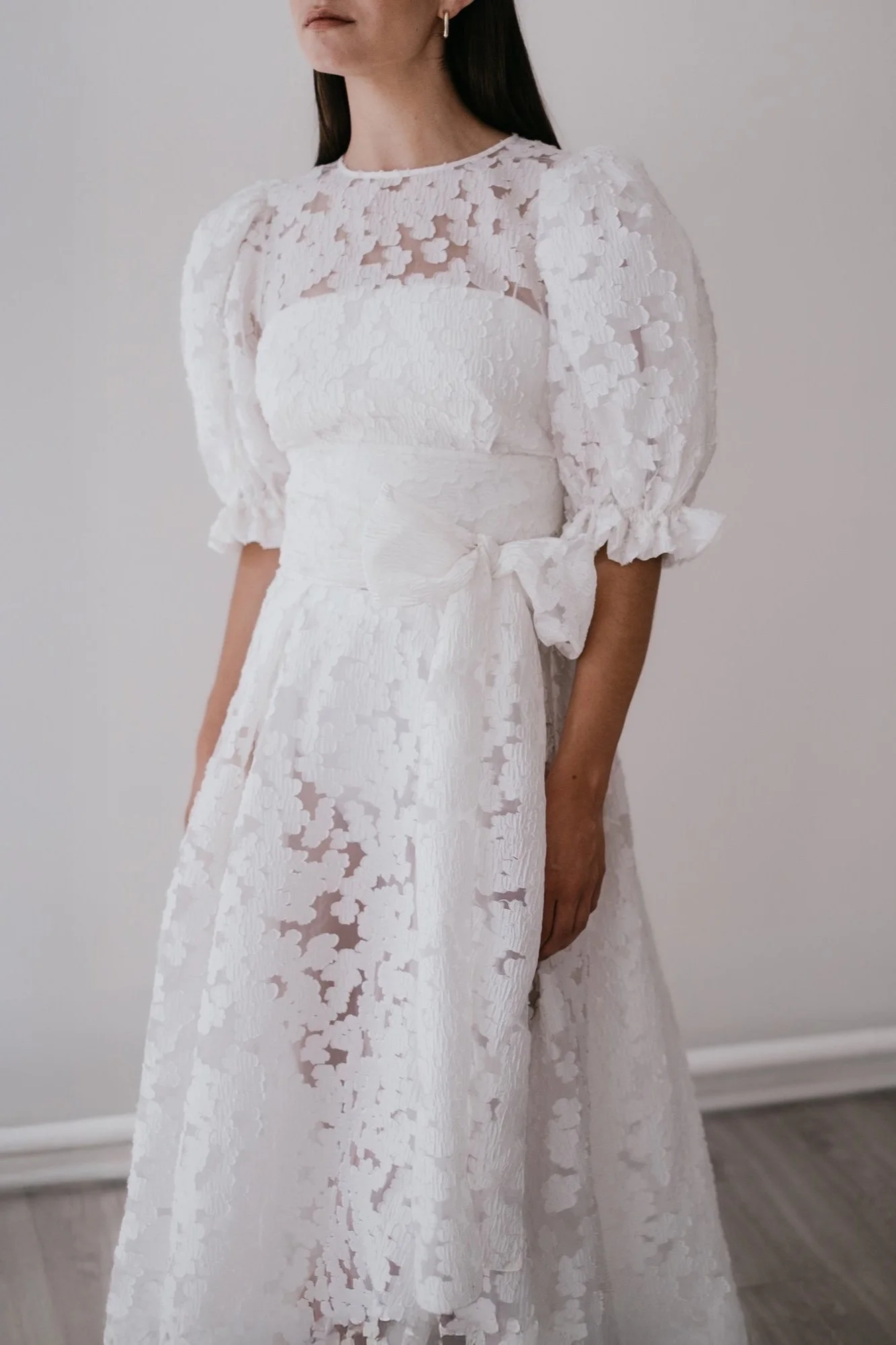 A woman wearing a white lace dress with puffed sleeves and a waist bow, standing against a plain background.