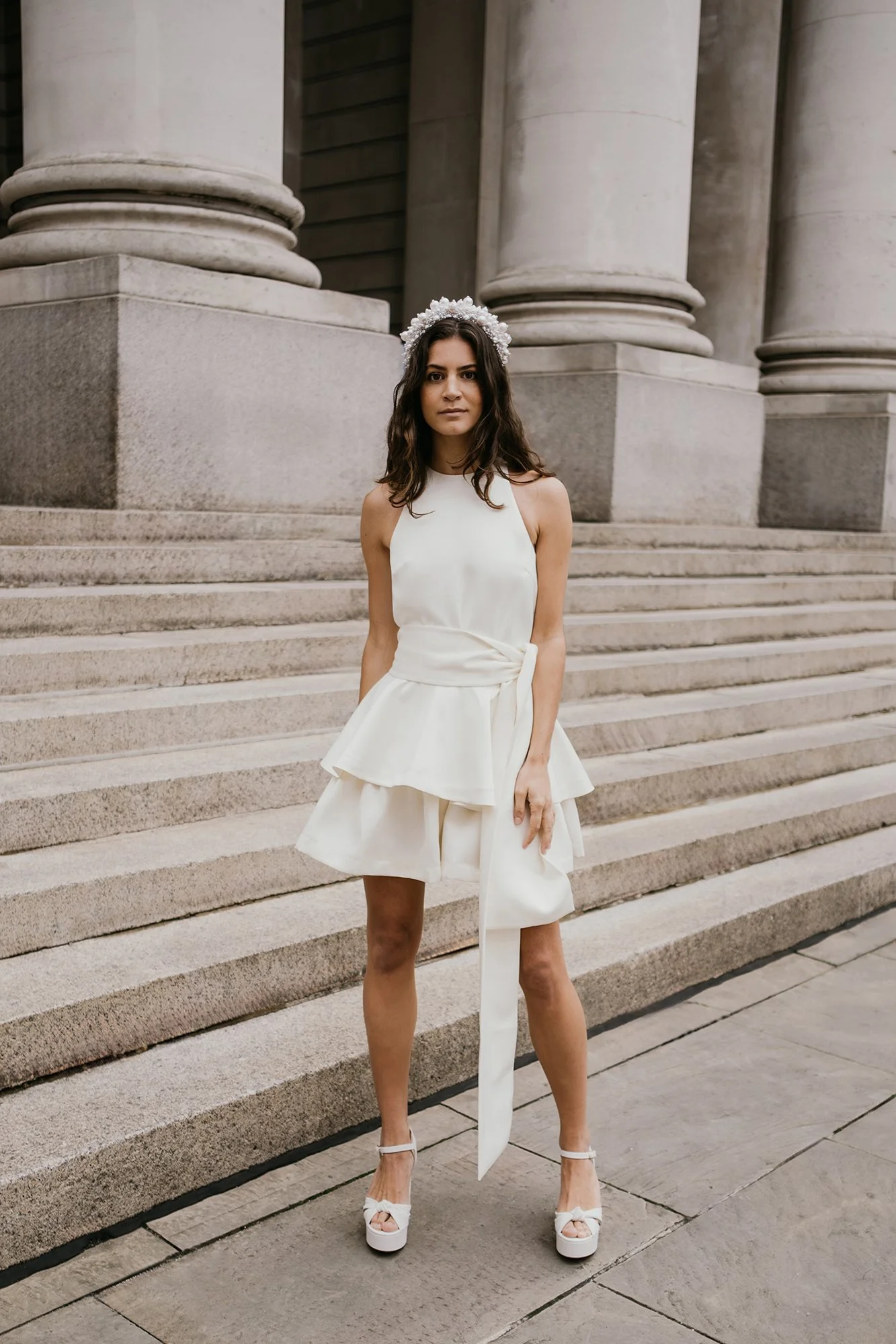 A young woman in a white dress and high heels standing on stone steps in front of large stone columns.