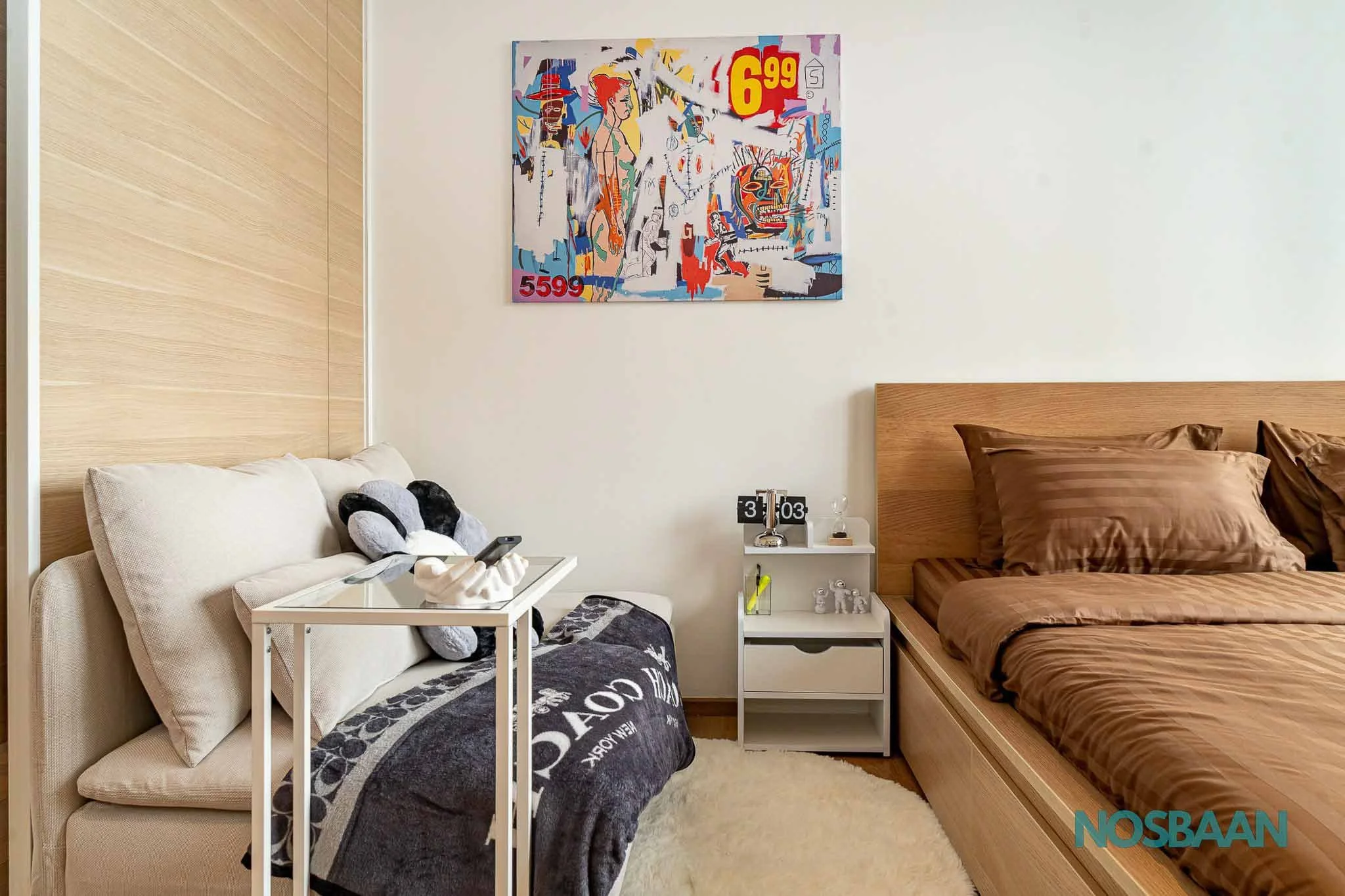 The image shows a bedroom with a beige sofa on the left, a small white side table beside the sofa, and a bed with brown bedding on the right. On the wall, there is a colorful abstract painting with various shapes and bright colors, including red and yellow. The nightstand has a digital clock, a glass, and some small objects. The wall is plain white, and the floor is covered with a light-colored rug.