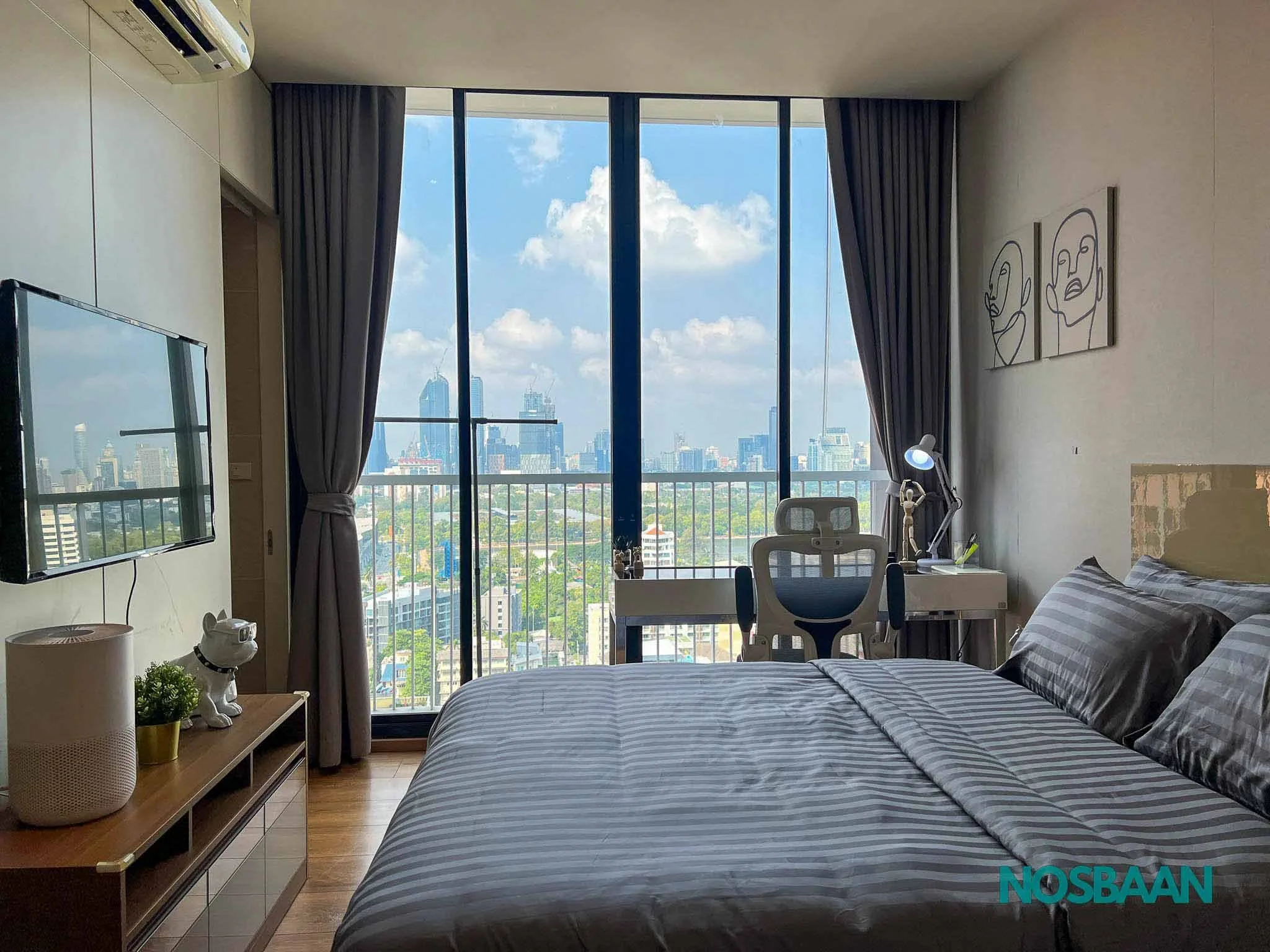 A modern bedroom with a large floor-to-ceiling window showing a city skyline with high-rise buildings and a partly cloudy sky. The room has a bed with gray striped bedding, a white desk with a chair, and wall art depicting abstract faces. Curtains frame the window, and a small table beside the bed holds decorative items including a dog statue and a plant.