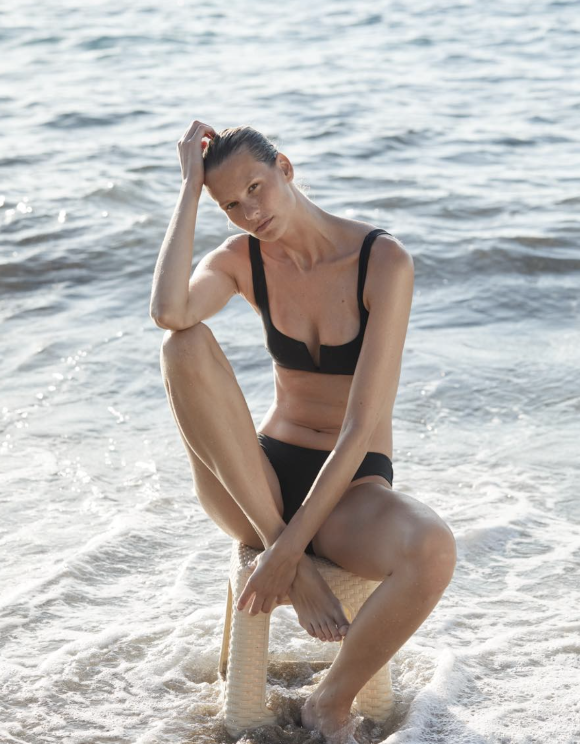 A woman in a black bikini sitting on a woven stool at the edge of the water on a beach, with her right hand in her hair and her left hand on her ankle, looking at the camera.