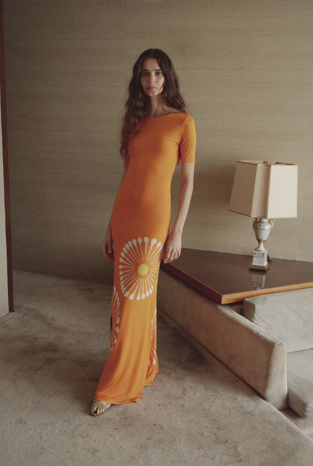 A woman with long brown hair wearing an orange dress with white floral patterns standing in a room with beige walls, a carpeted floor, a wooden side table, and a white table lamp.