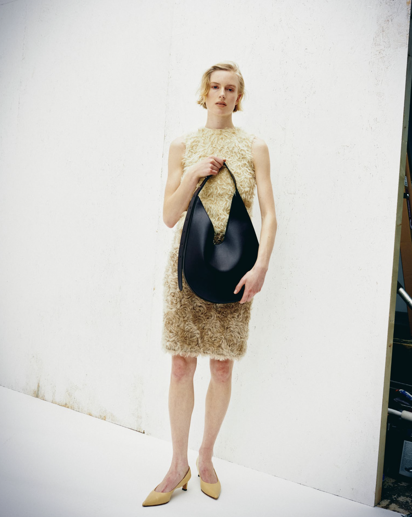 A woman with short blonde hair wearing a sleeveless, knee-length, curly beige dress and yellow high heels, holding a large black handbag, standing against a plain white wall.