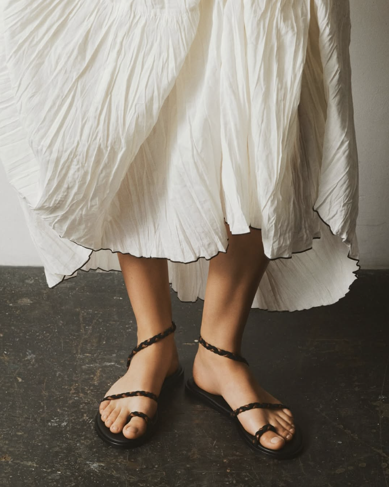 Person wearing a flowing, pleated, white skirt and black sandals with braided straps, standing on a dark floor.