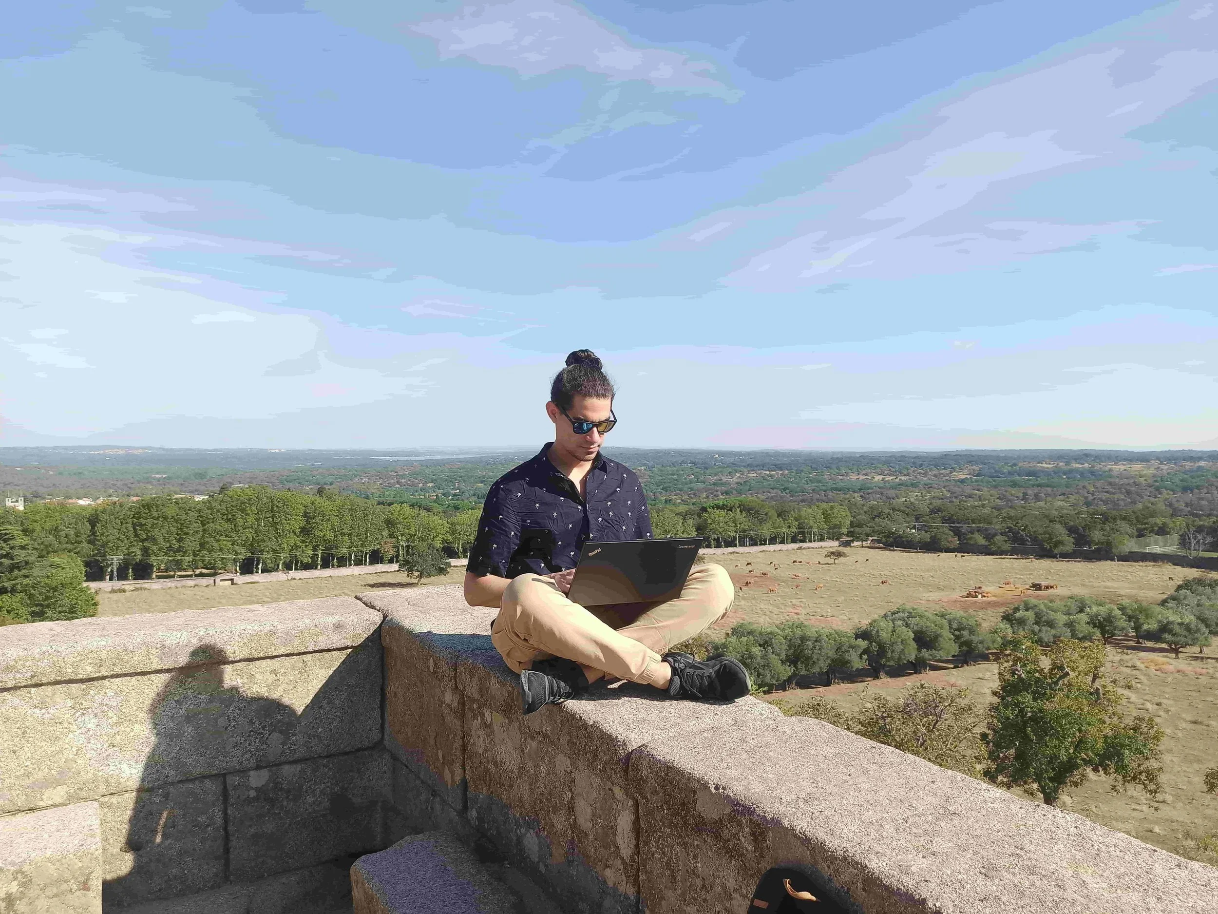 A guy sitting on a wall with a laptop with nature in the background