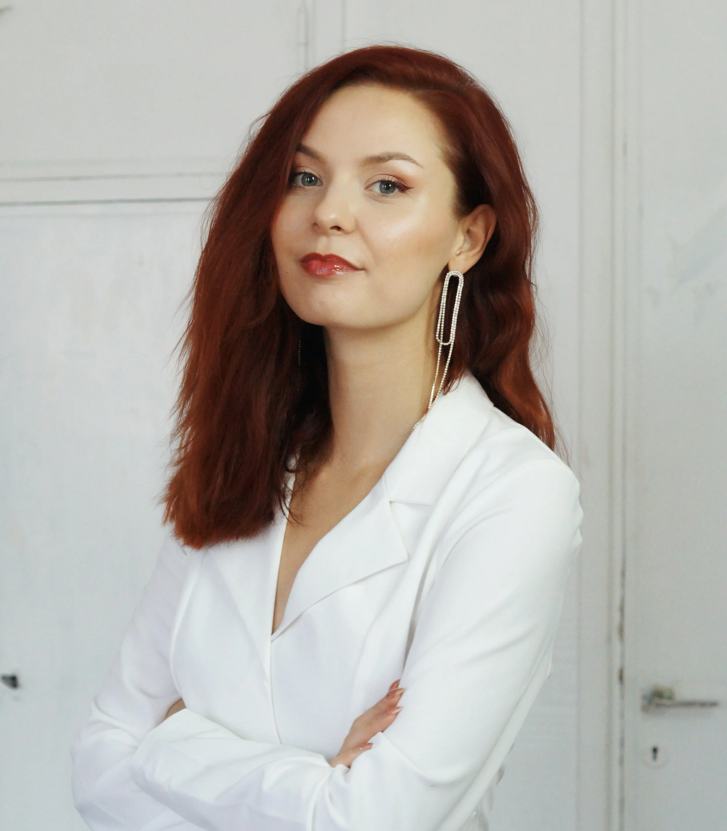 Promdress nainen, jossa punaiset hiukset, valkoisessa yläosassa, kerroksellinen korvakoru, käsivarret ristissä, tausta valkoinen seinä.