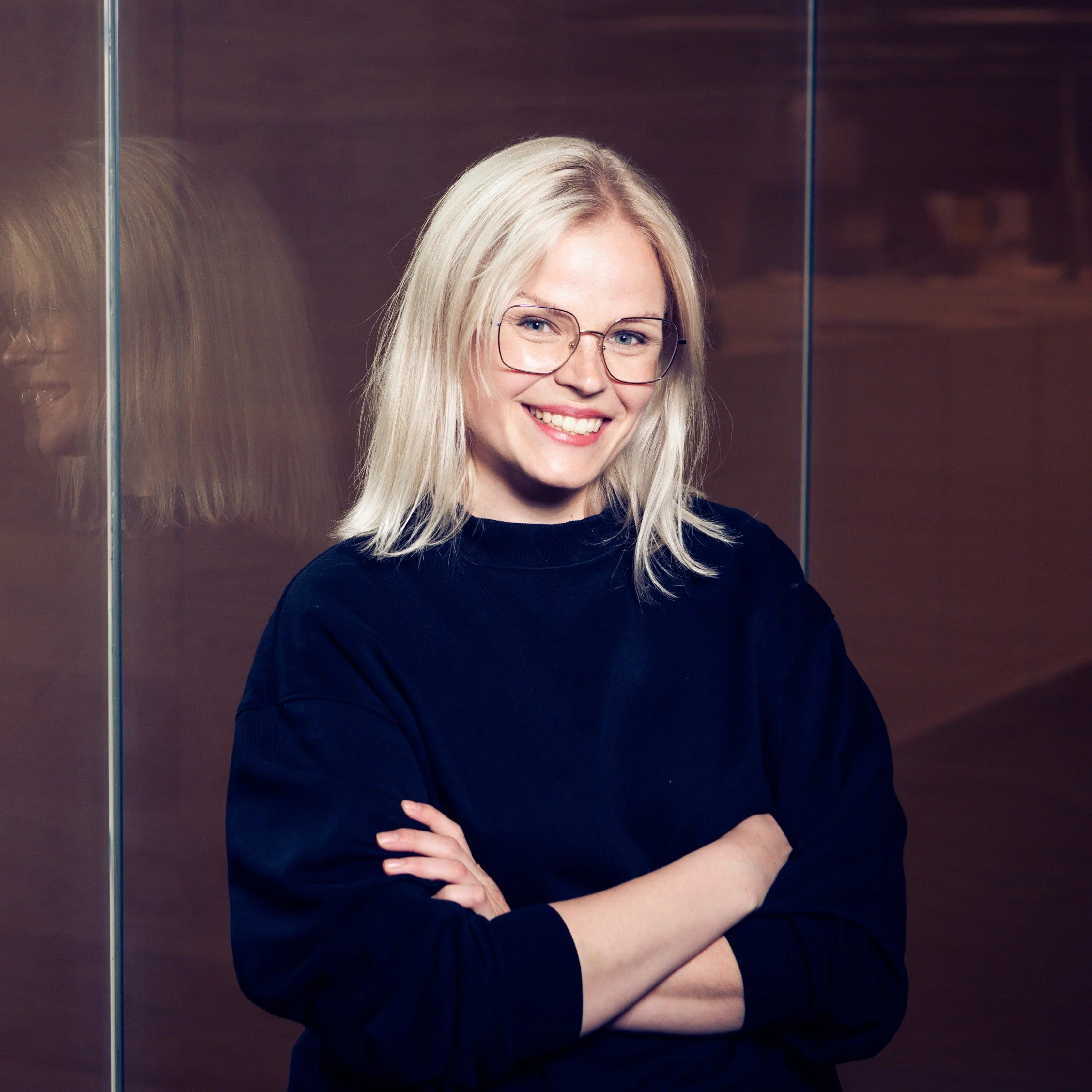 Hymyilevä nuori nainen, jolla on vaaleat hiukset ja silmälasit, poseeraa sisätiloissa.