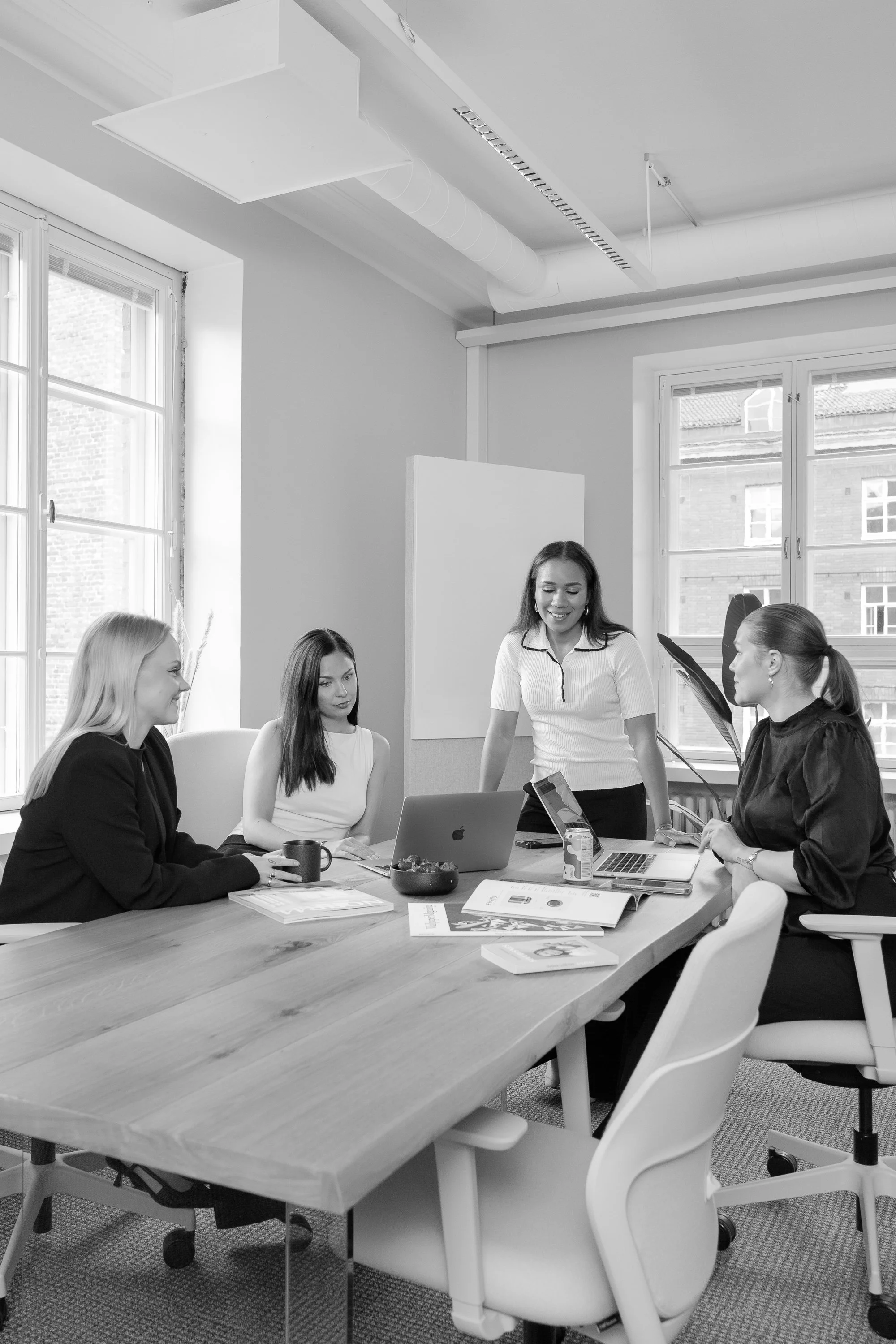Four women having a meeting in a modern office, with laptops, notebooks, and coffee mugs on the table.