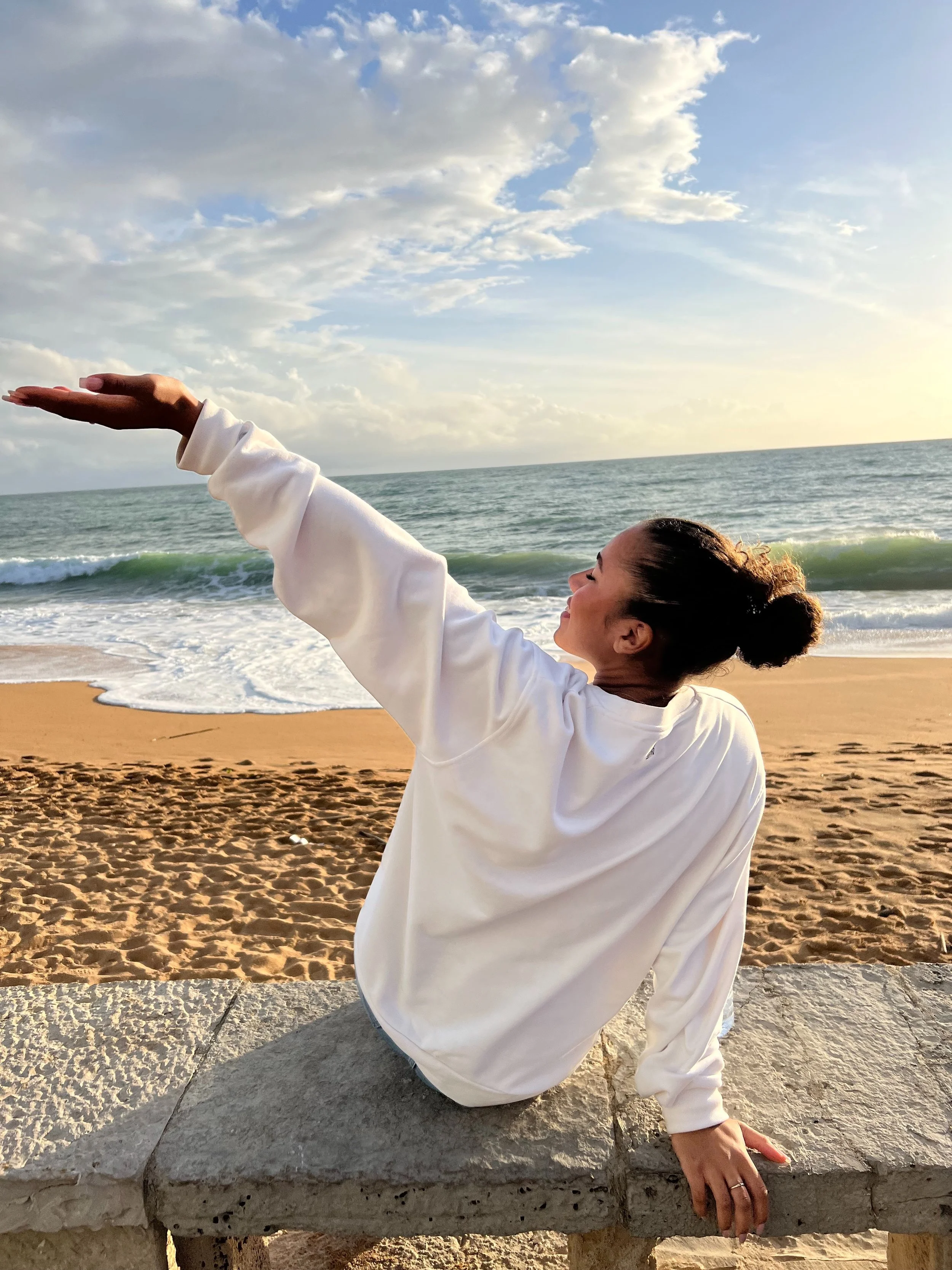 Nainen istuu kivijalustalla rannalla, nostaa käsiään kohti taivasta auringonlaskun aikaan.