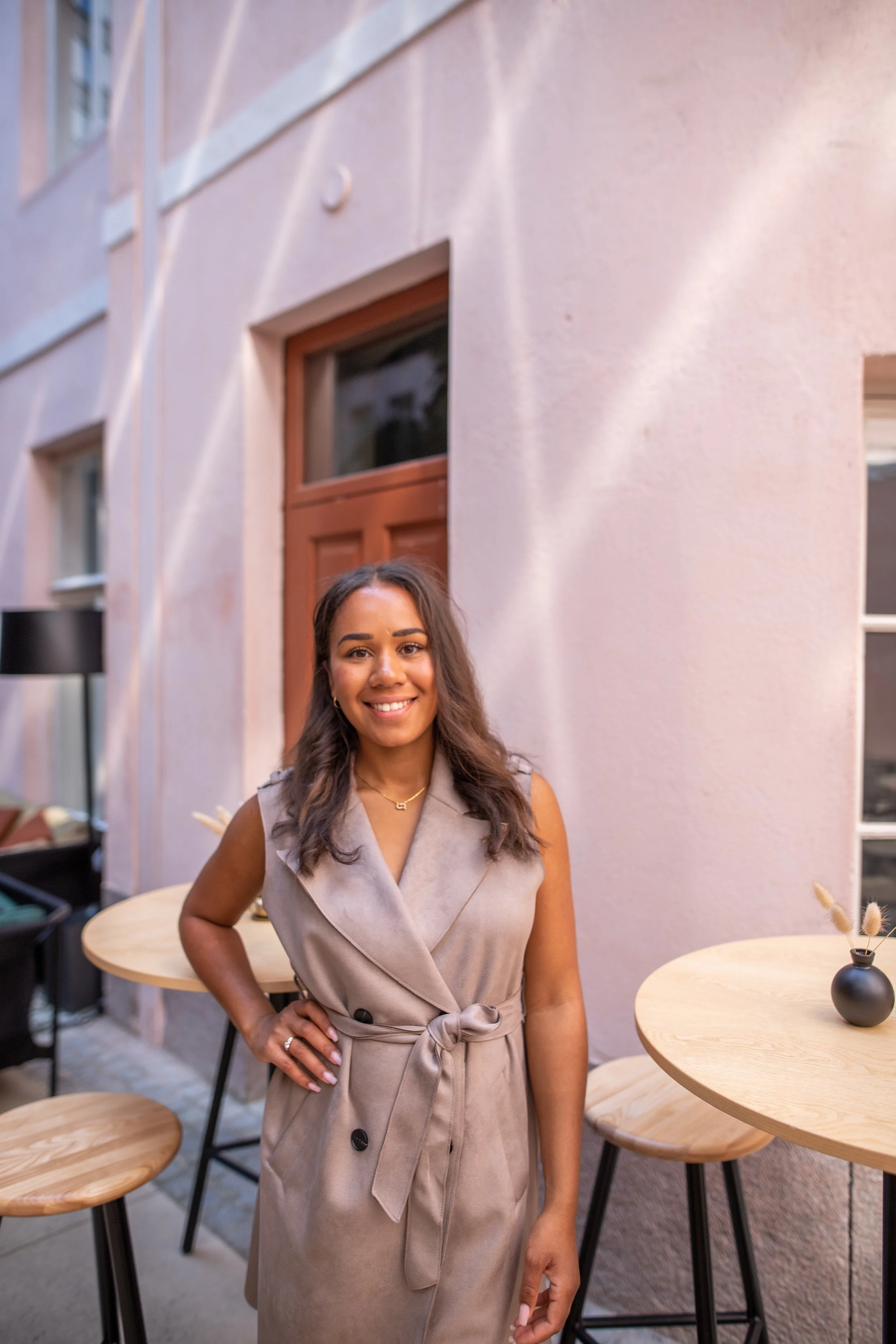 Nainen hymyilemässä istuu kahvilassa, taustalla vaaleanpunainen seinä ja pieniä pöytiä ja kukkavazoja.