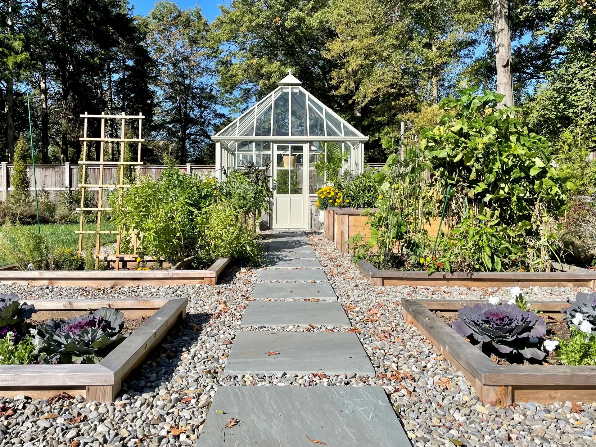 A garden pathway leading to a glass greenhouse, surrounded by vegetable beds and flowering plants, with trees in the background and clear blue sky.