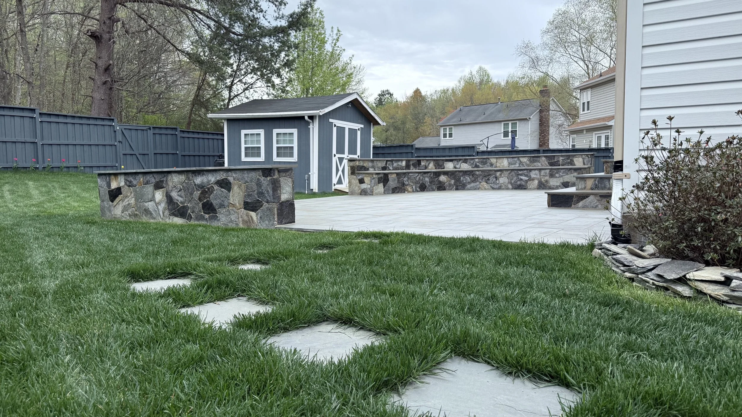 View of a backyard with a grassy lawn, a stone patio, a small blue shed with white trim, a dark blue fence, and neighboring houses in the background.