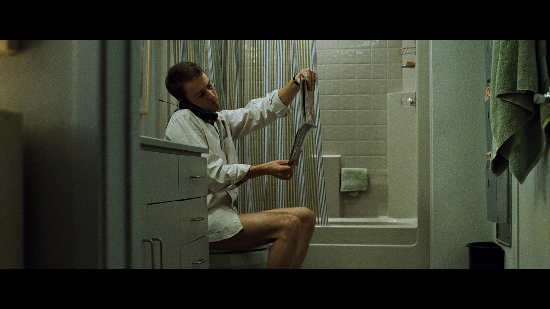 Man on toilet talking on phone reading magazine in bathroom