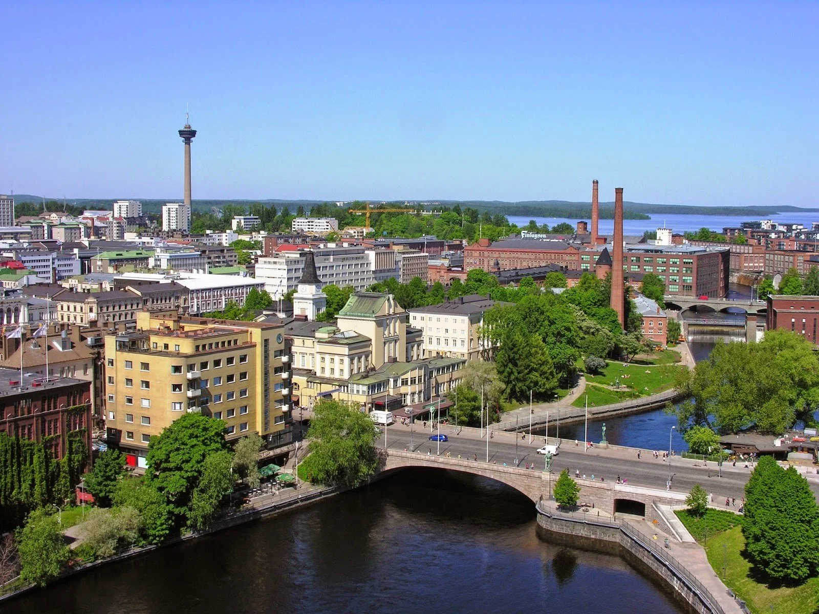 Aerial view of Tampere, Finland, showcasing Näsinneula tower, red brick industrial buildings, and green parks by a river.