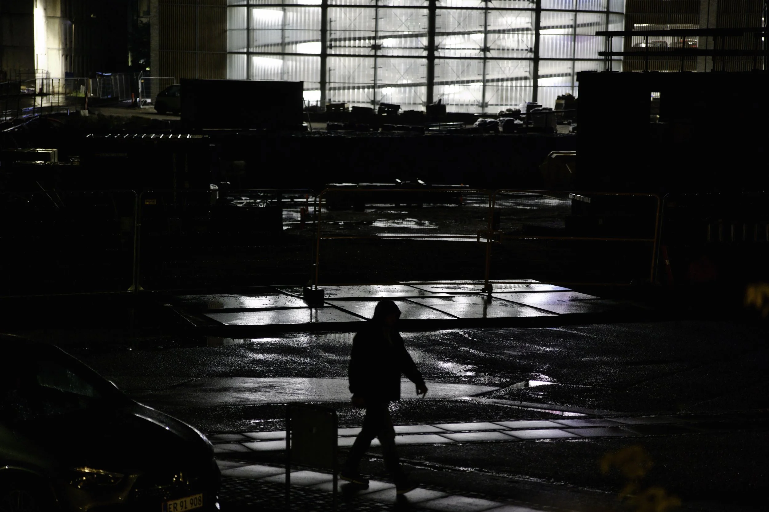 Pedestrian in the rain against a backdrop of a construction site and bright lights from a parkade - Carlsensvej, Køge, Denmark