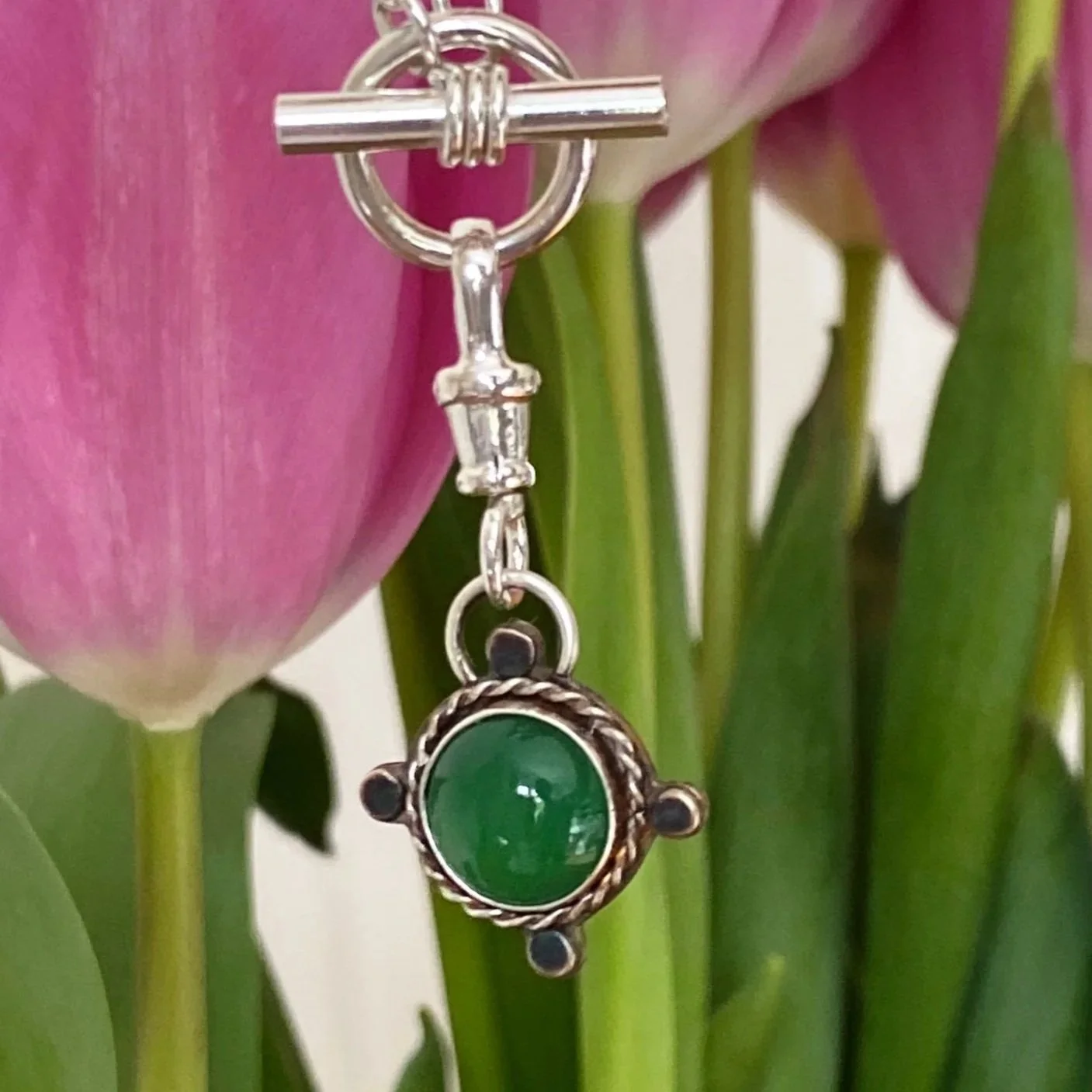 Walter and Nancy Jewellery's handmade, sterling silver and green onyx necklace charm hanging in front of pink tulips..